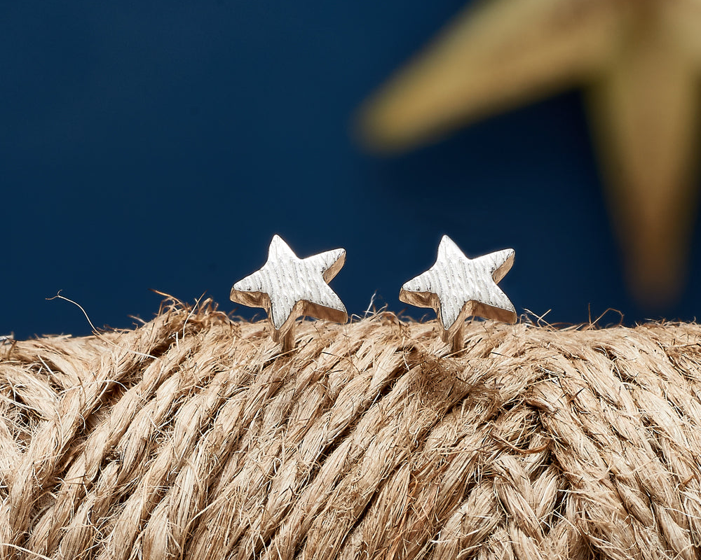 A pair of small star-shaped stud earrings with a textured silver finish, displayed on a bundle of natural jute rope against a dark blue background with a blurred star motif.