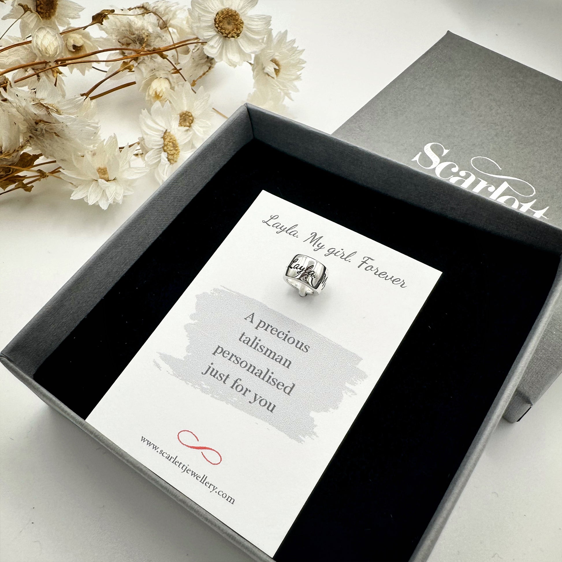 Jewelry box with a card featuring a silver charm and branding on a white background with flowers.