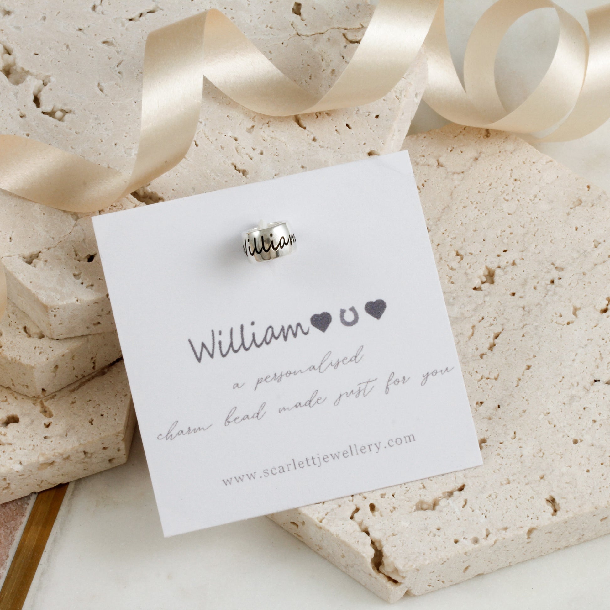 Silver charm bead on a card with 'William' engraved, displayed on a textured stone surface with ribbons.