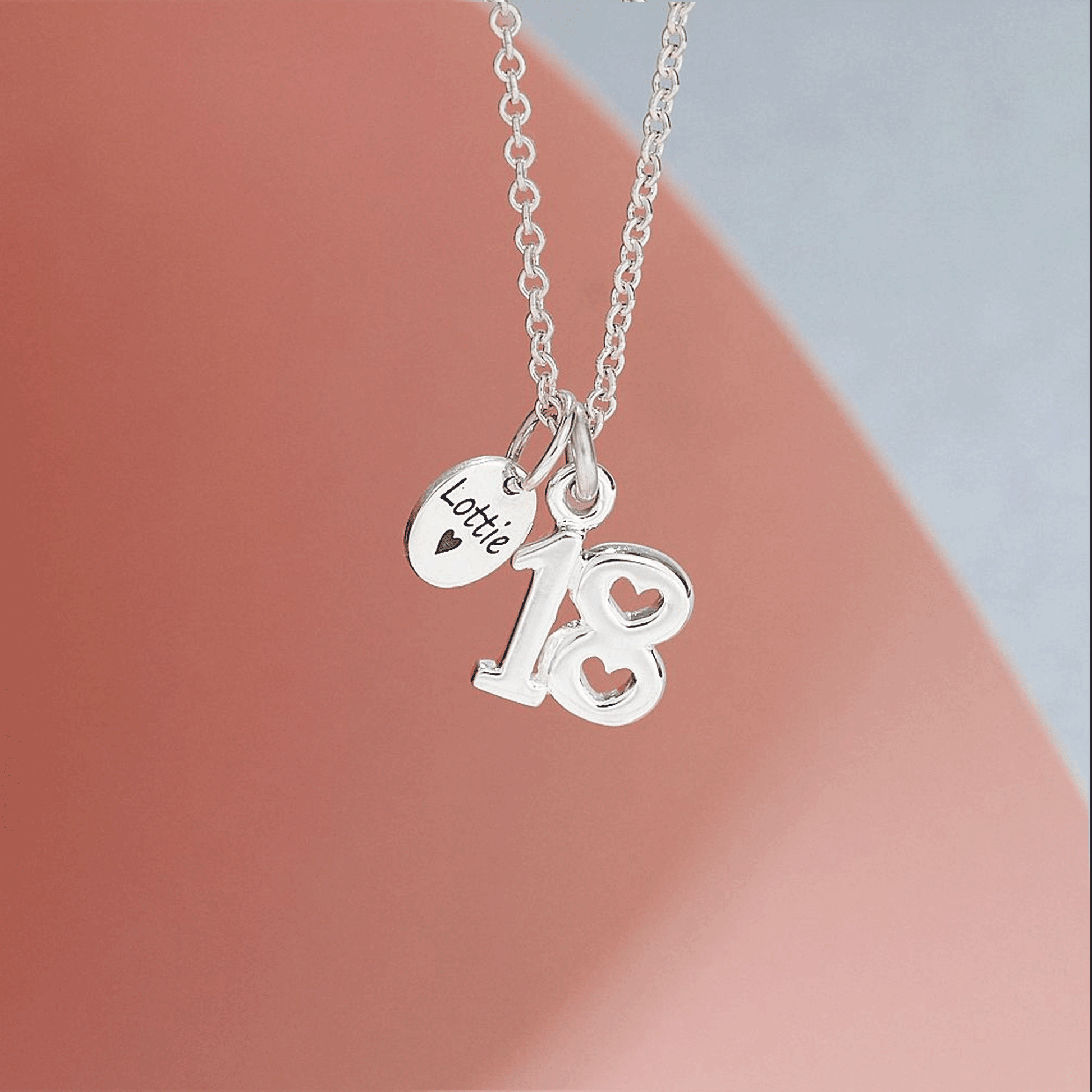 Silver necklace with '18' and 'Lottie' charms on a soft pink and gray background