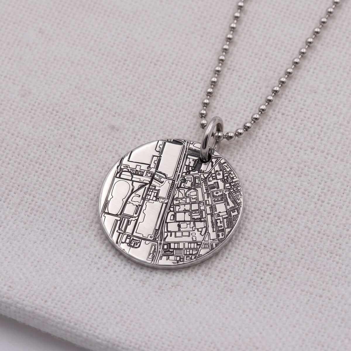 A silver round pendant necklace featuring an engraved city map design, suspended from a silver ball chain on a textured white fabric background.