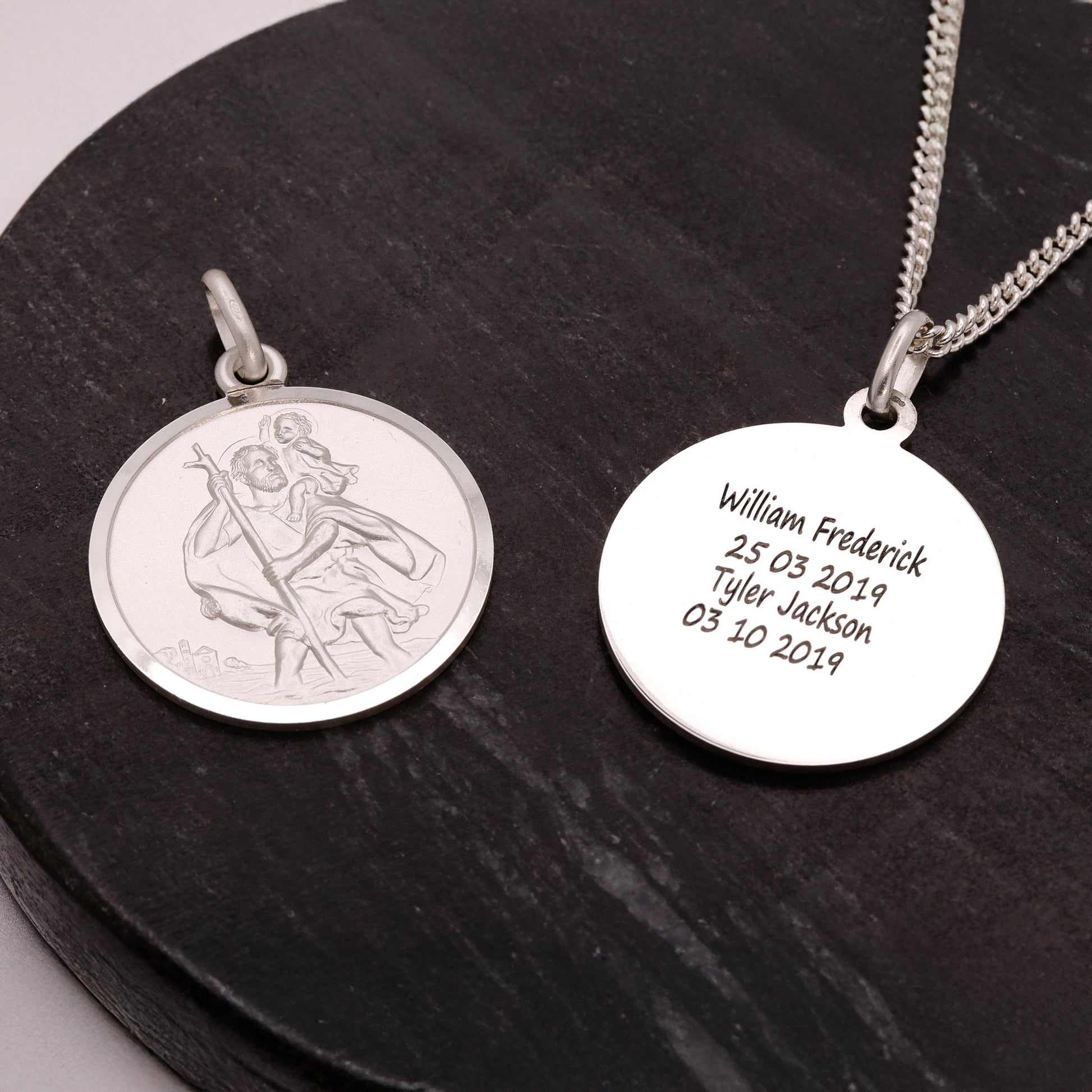 Two silver pendants on a dark surface, one with an engraved design and the other with engraved text.