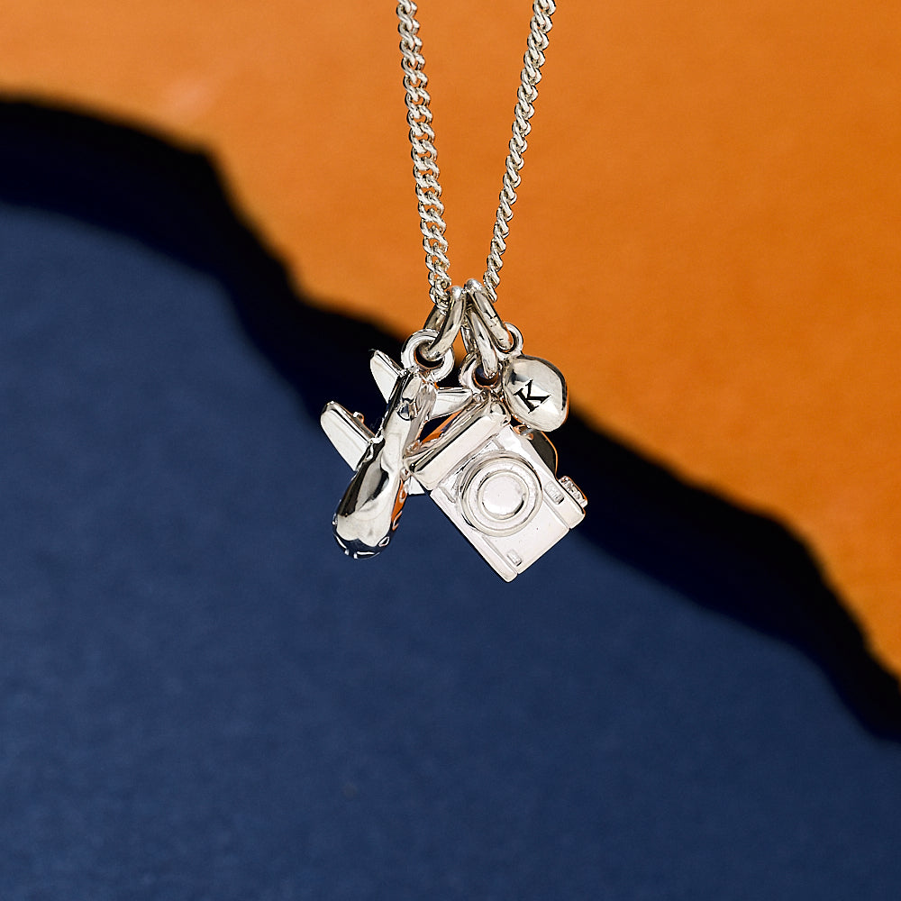 Silver necklace with camera charm and 'K' pendant on a blue and orange gradient background