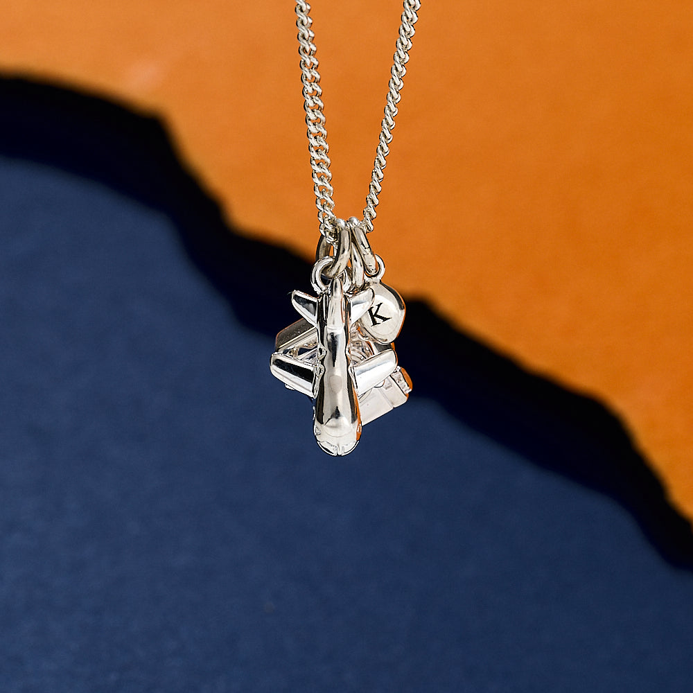 Silver necklace with three charms, camera aeroplane and pebble, on a blue and orange gradient background