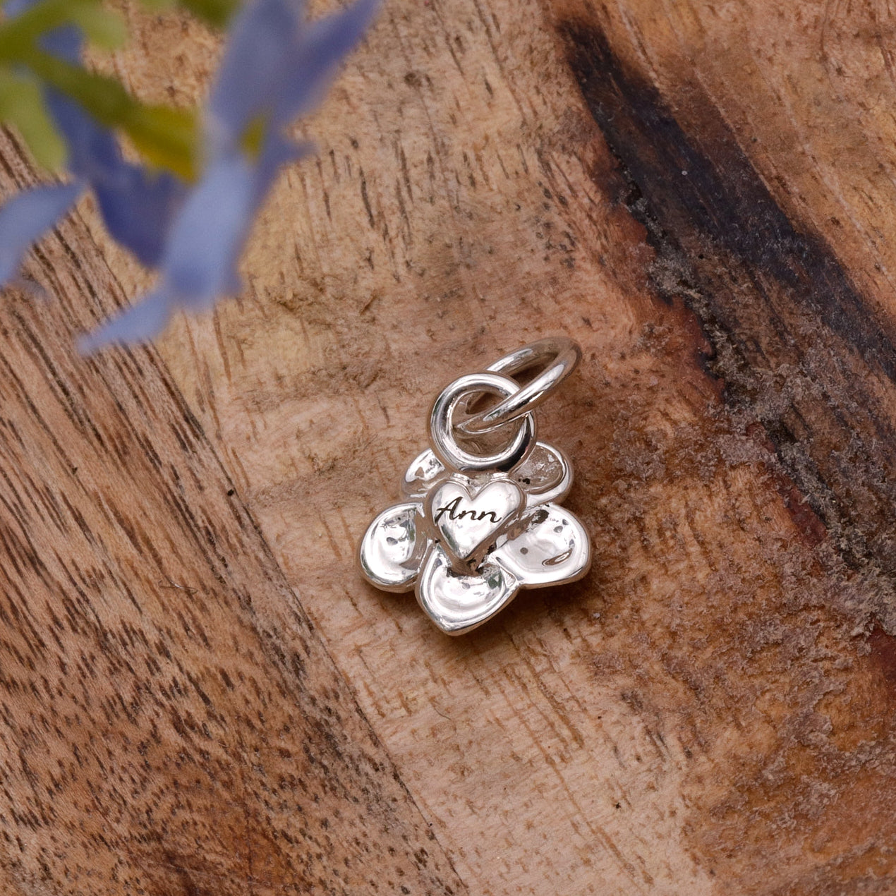 Silver charm with 'Ann' engraved on a wooden surface with blue flowers.