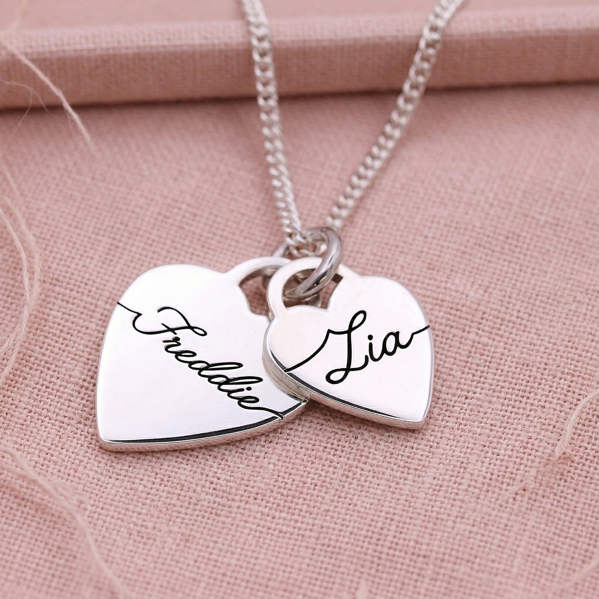 Two heart-shaped pendants on a chain with names engraved on a pink fabric background