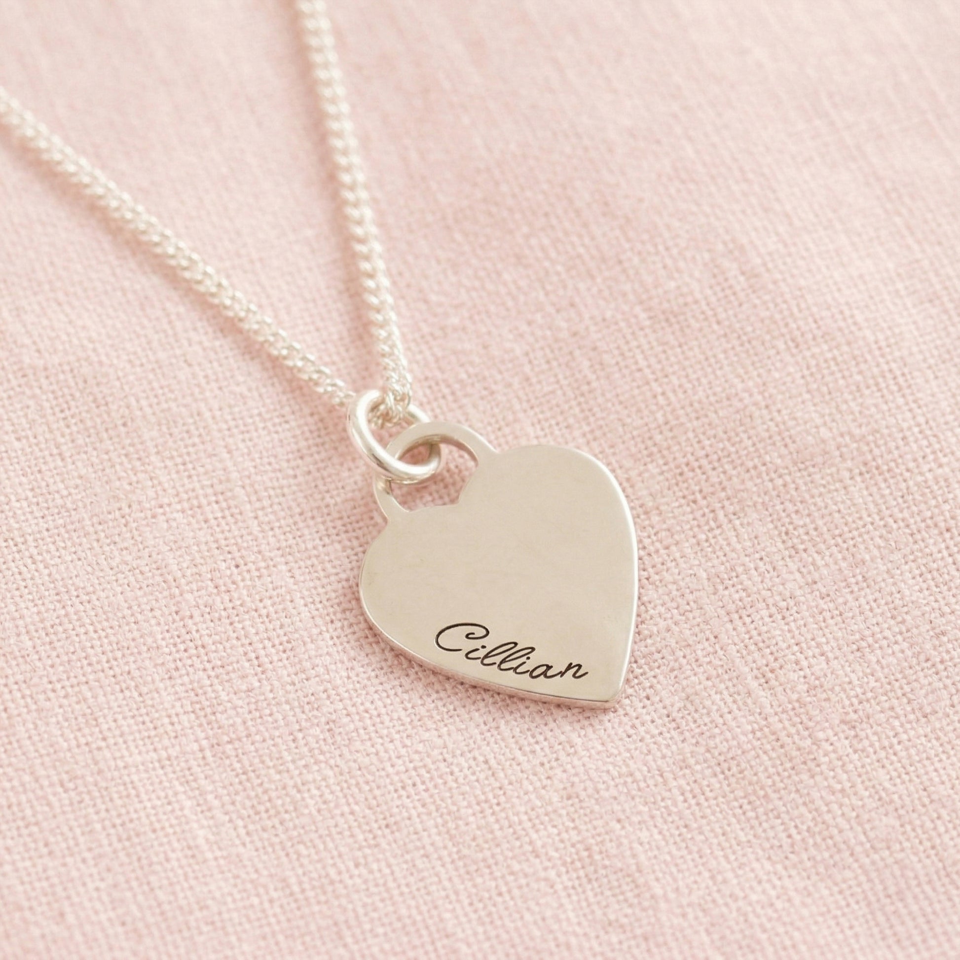 Silver heart-shaped pendant necklace with engraved name on a pink fabric background