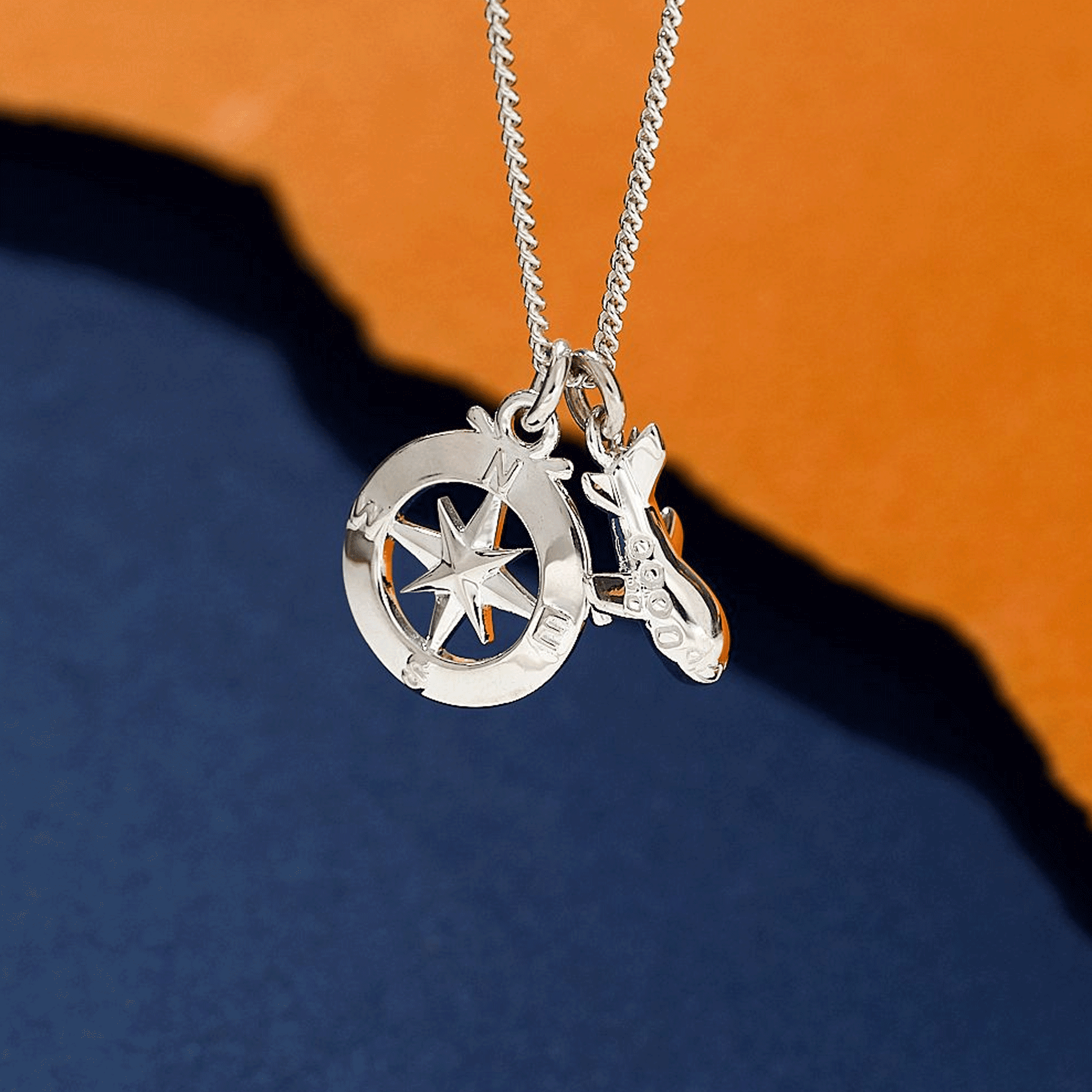 Silver necklace with a aeroplane and compass pendant on an orange and blue gradient background