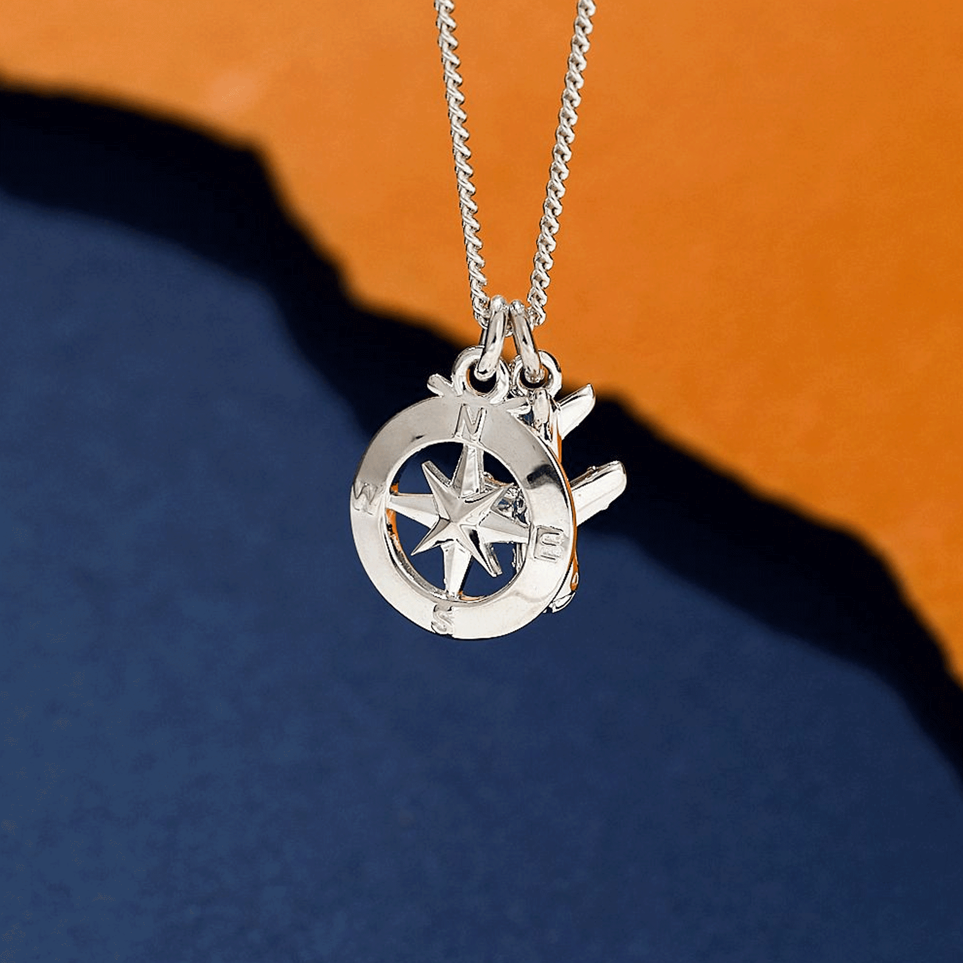 Silver necklace with a aeroplane and compass pendant on an orange and blue gradient background