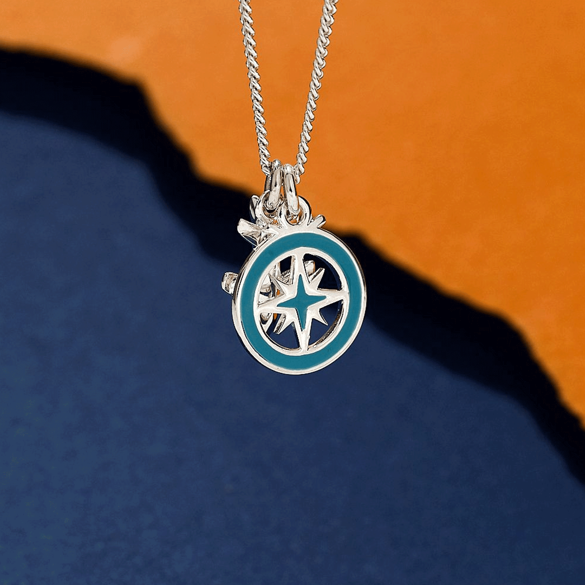 Silver necklace with a aeroplane and compass pendant on an orange and blue gradient background