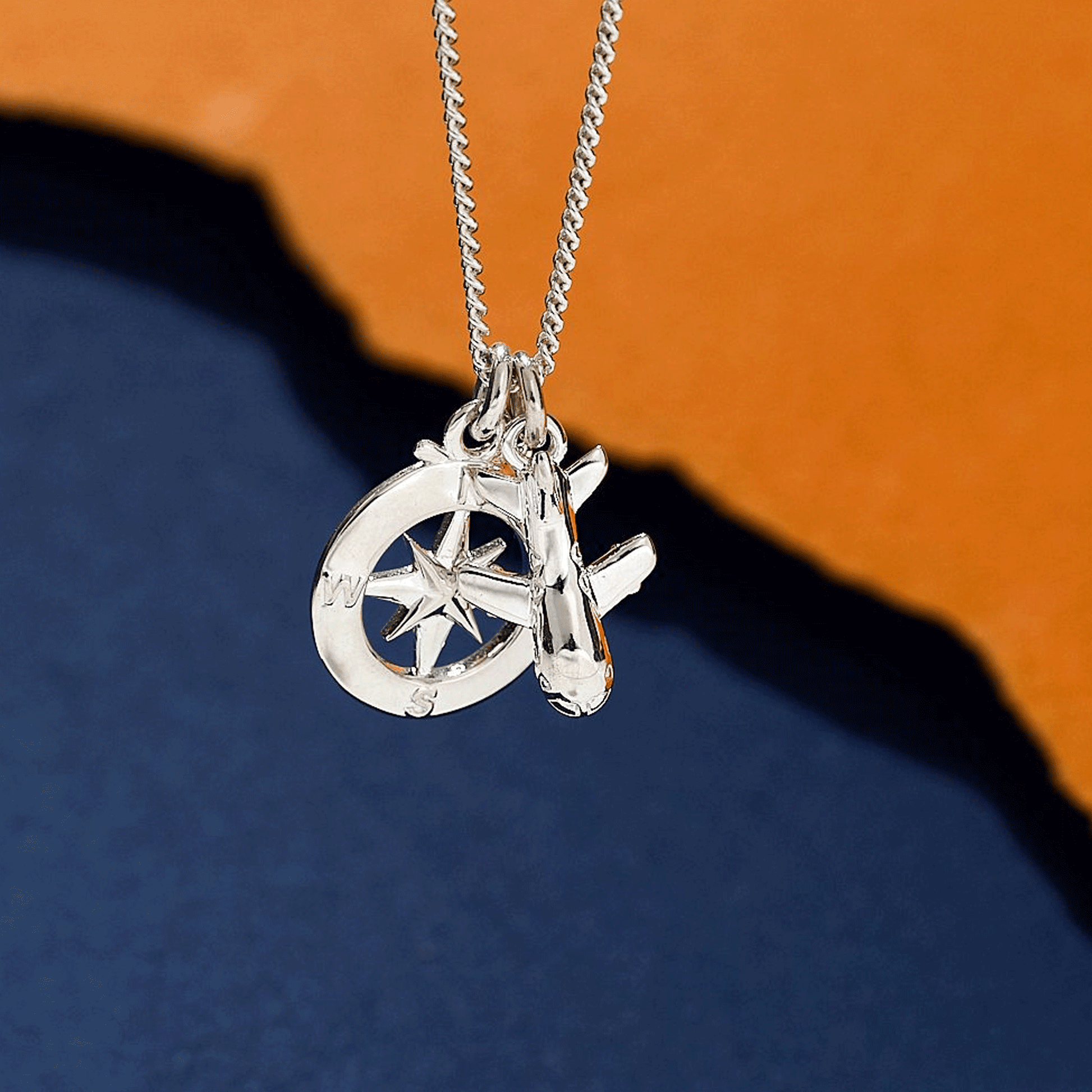 Silver necklace with a aeroplane and compass pendant on an orange and blue gradient background