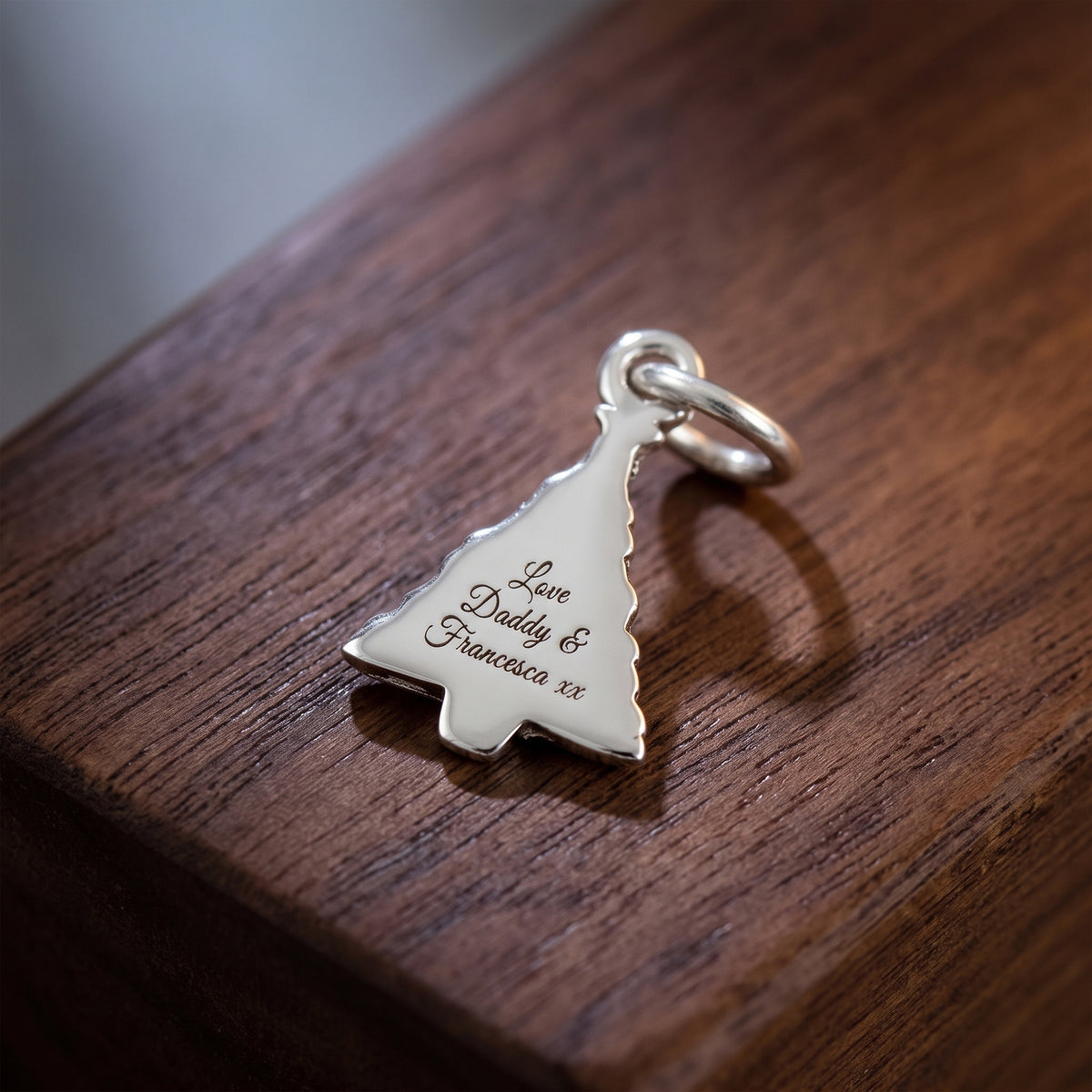 A silver Christmas tree-shaped pendant engraved with 'Love Daddy & Francesca XX' resting on a dark brown wooden surface.