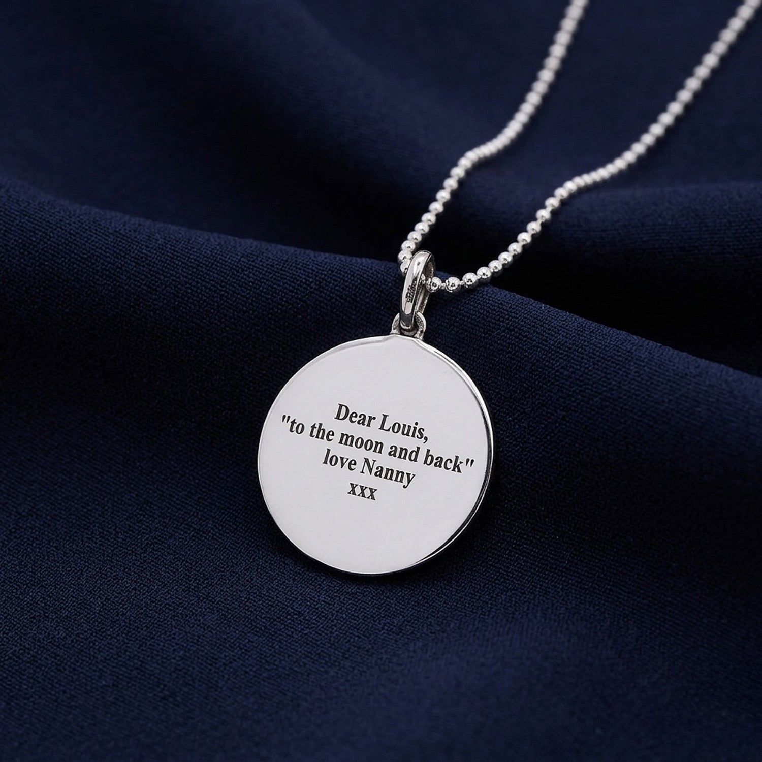 A silver round pendant necklace with a ball chain, featuring engraved text that reads 'Dear Louis, "to the moon and back" love Nanny xxx', displayed on a dark navy blue fabric background.