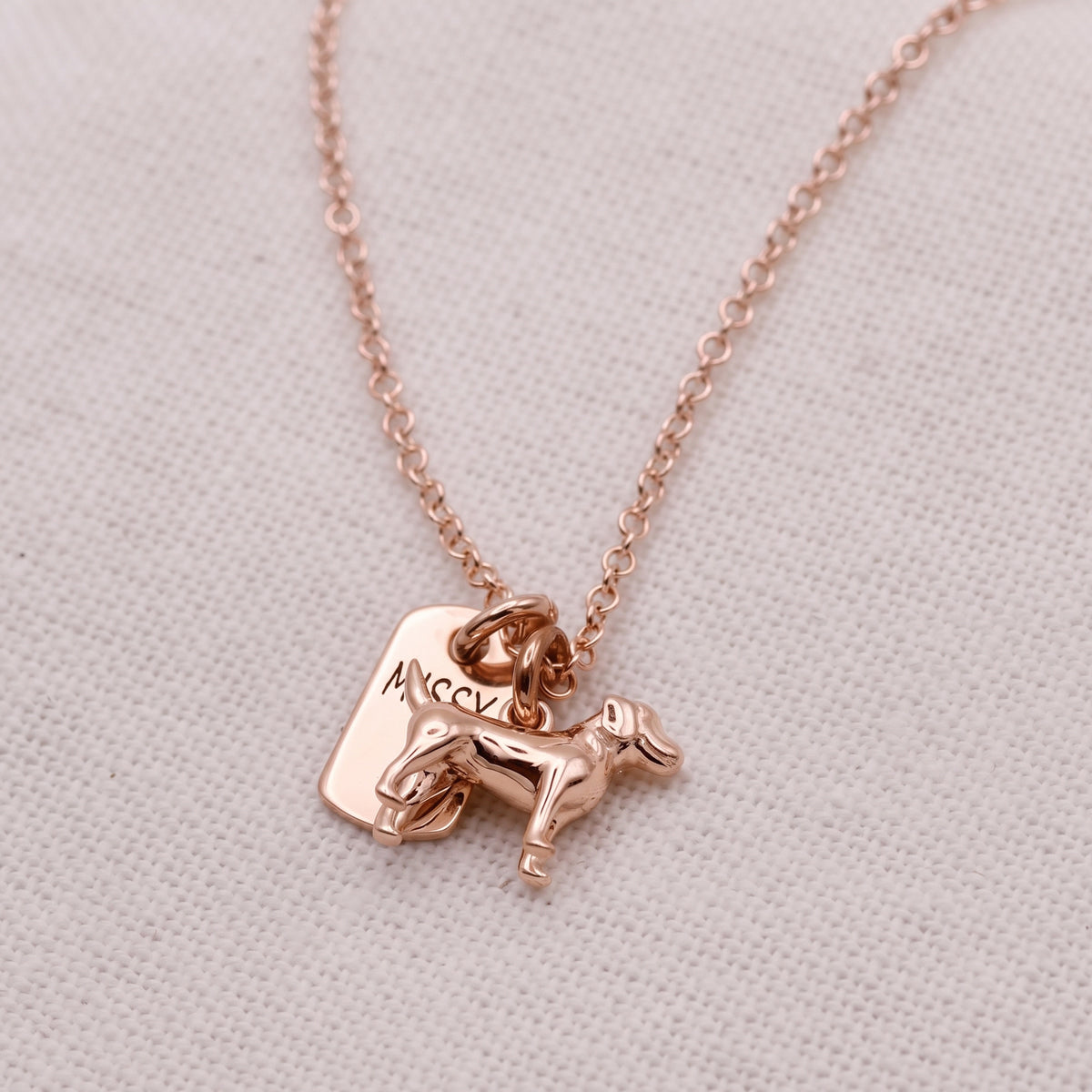 A rose gold necklace with a small dog charm and a rectangular tag engraved with the name 'Maggie', displayed on a textured beige fabric background.