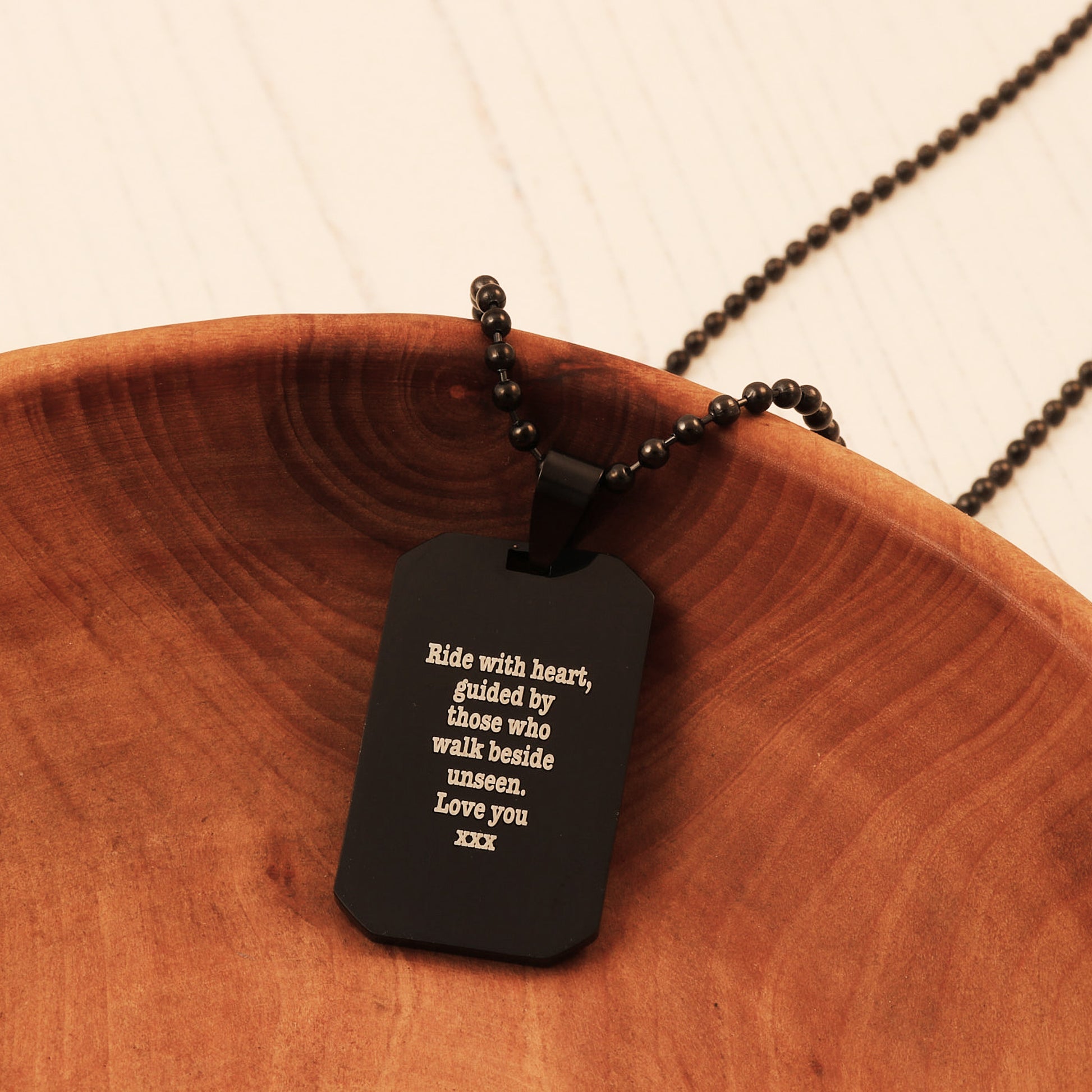 Black dog tag pendant with engraved text on a wooden surface