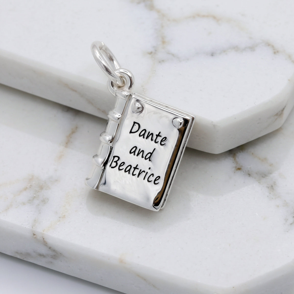 A silver book-shaped pendant charm engraved with the words 'Dante and Beatrice' in black script, resting on a white marble surface.