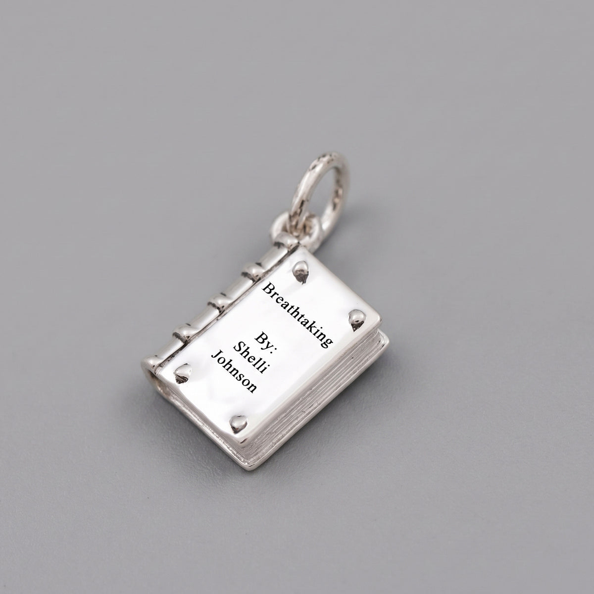 A silver book-shaped pendant charm engraved with the text 'Breathtaking By: Shelli Johnson', displayed on a neutral gray background.