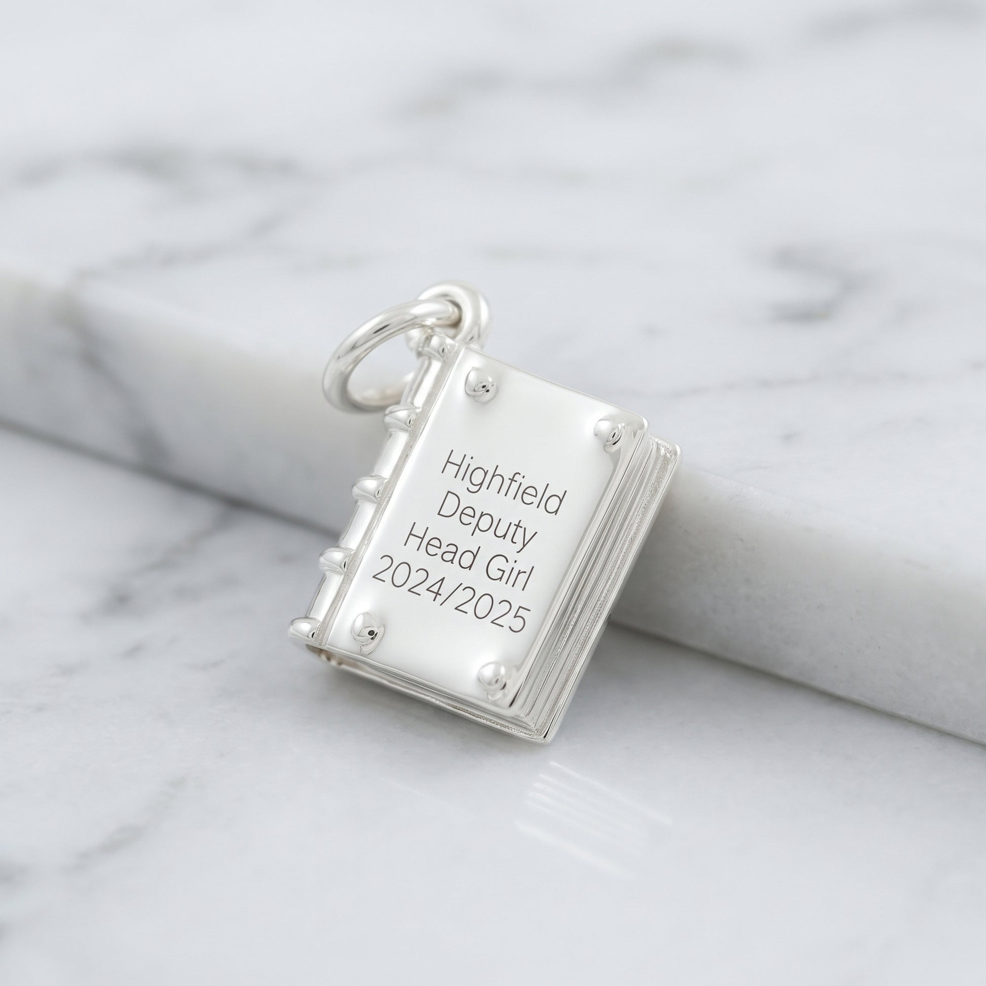 A silver book-shaped pendant charm engraved with 'Highfield Deputy Head Girl 2024/2025' resting on a white marble surface.