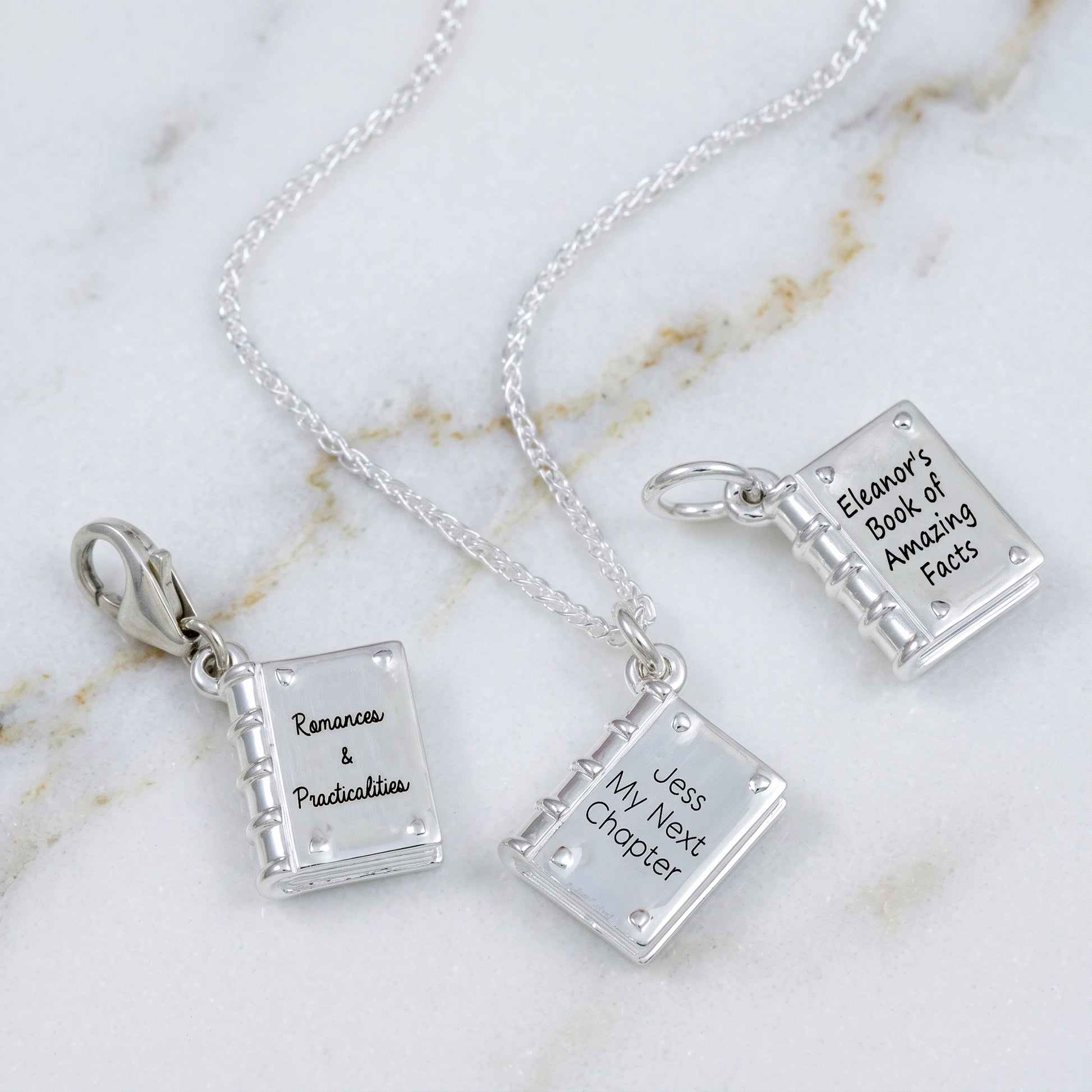 Three silver book-shaped pendant necklaces with engraved text, displayed on a white marble background. The pendants are shaped like open books and feature silver clasps.
