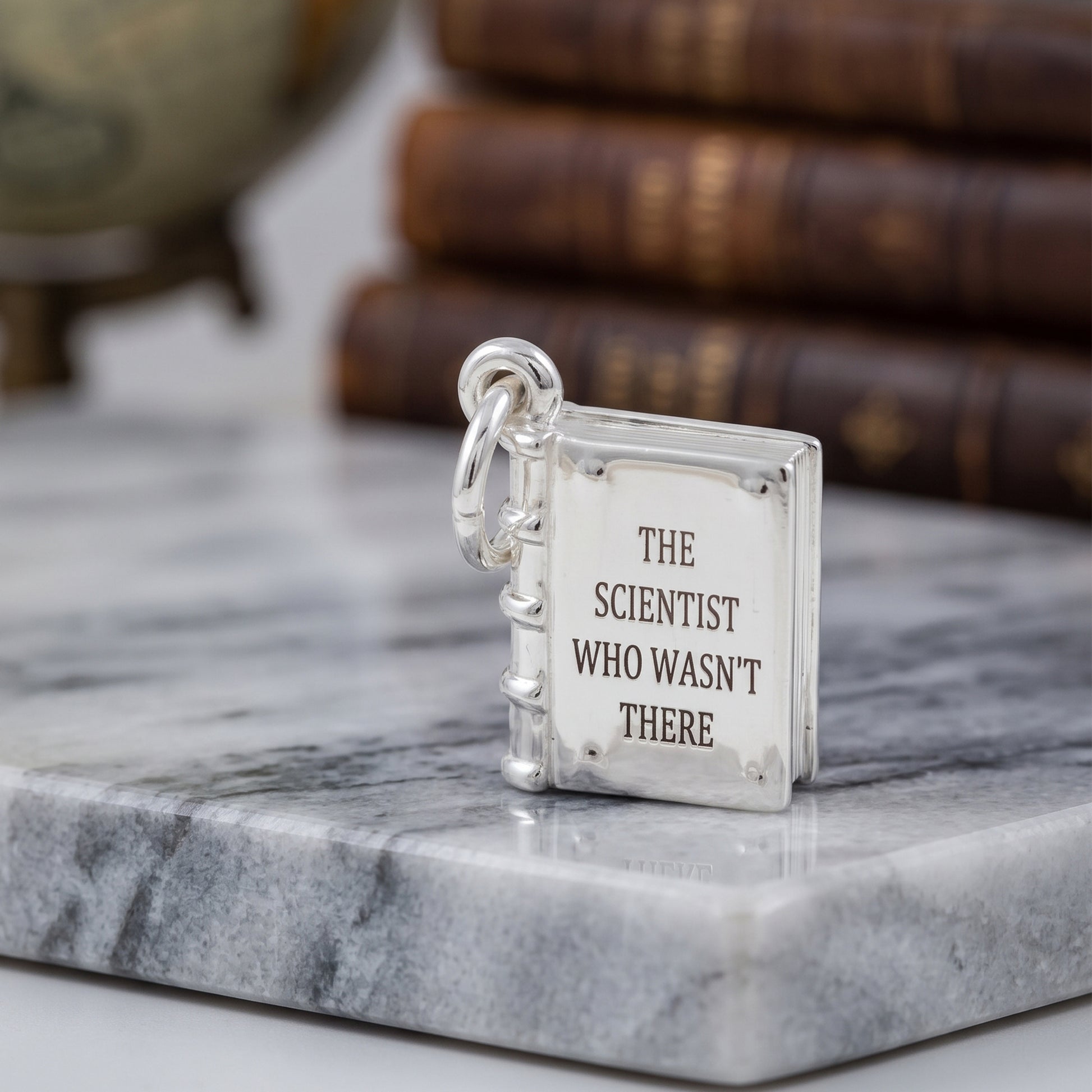 A silver book-shaped pendant charm with the engraved text 'THE SCIENTIST WHO WASN'T THERE' on its cover, resting on a gray marble slab with a blurred background of old books.