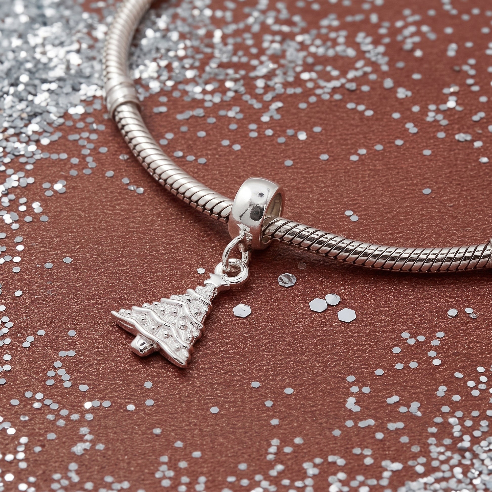 A silver snake chain bracelet with a dangling Christmas tree charm, displayed on a textured brown surface scattered with silver glitter.