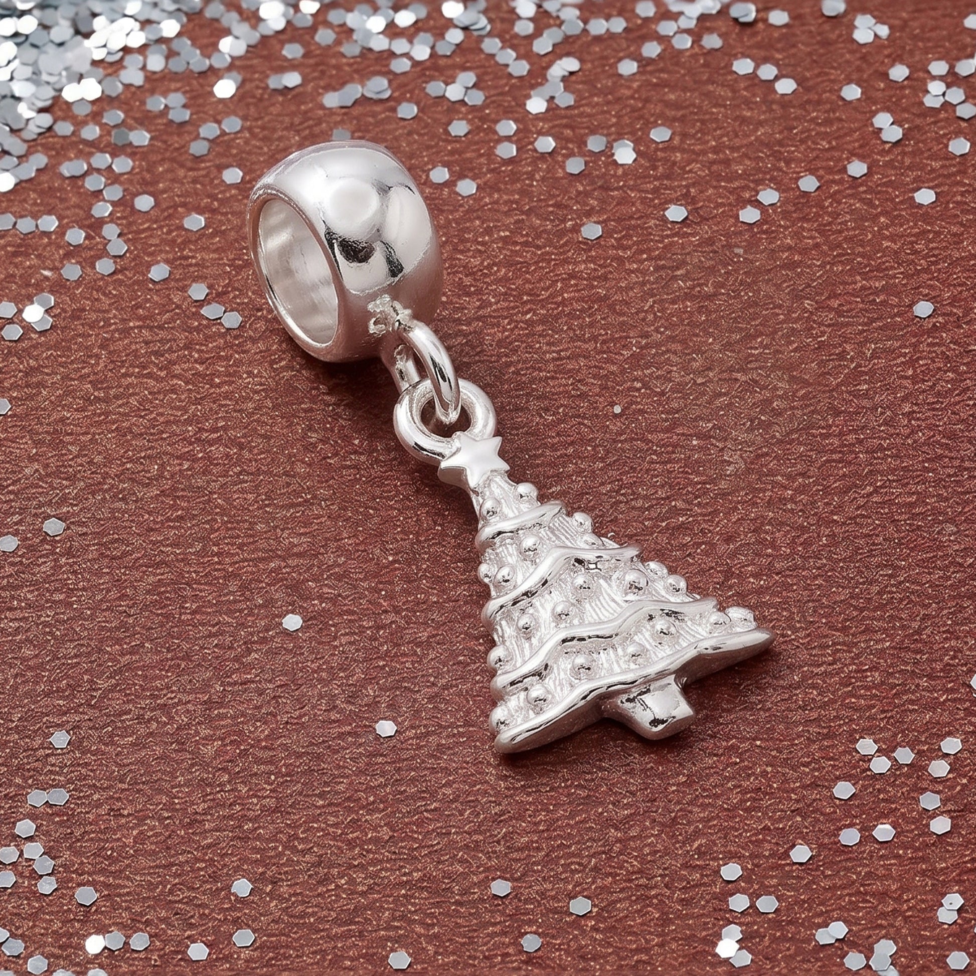 A silver Christmas tree charm bead with a polished dome top, displayed on a textured brown background with scattered silver glitter.