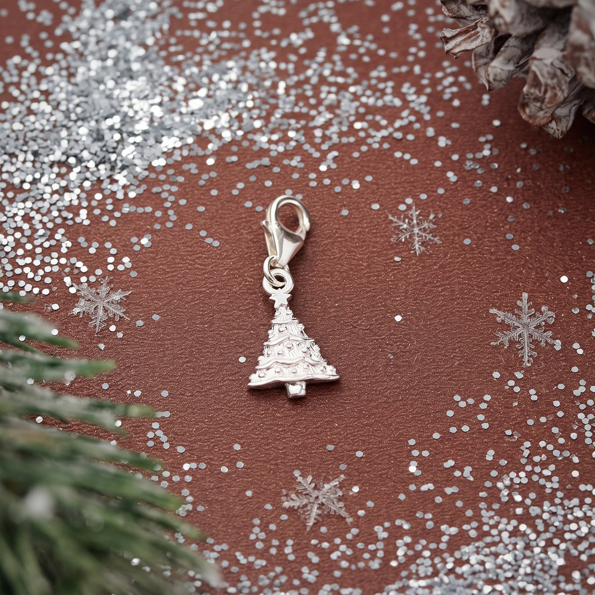 A silver Christmas tree charm with a lobster clasp, displayed on a textured brown surface decorated with silver glitter, snowflake cutouts, and pine branches.