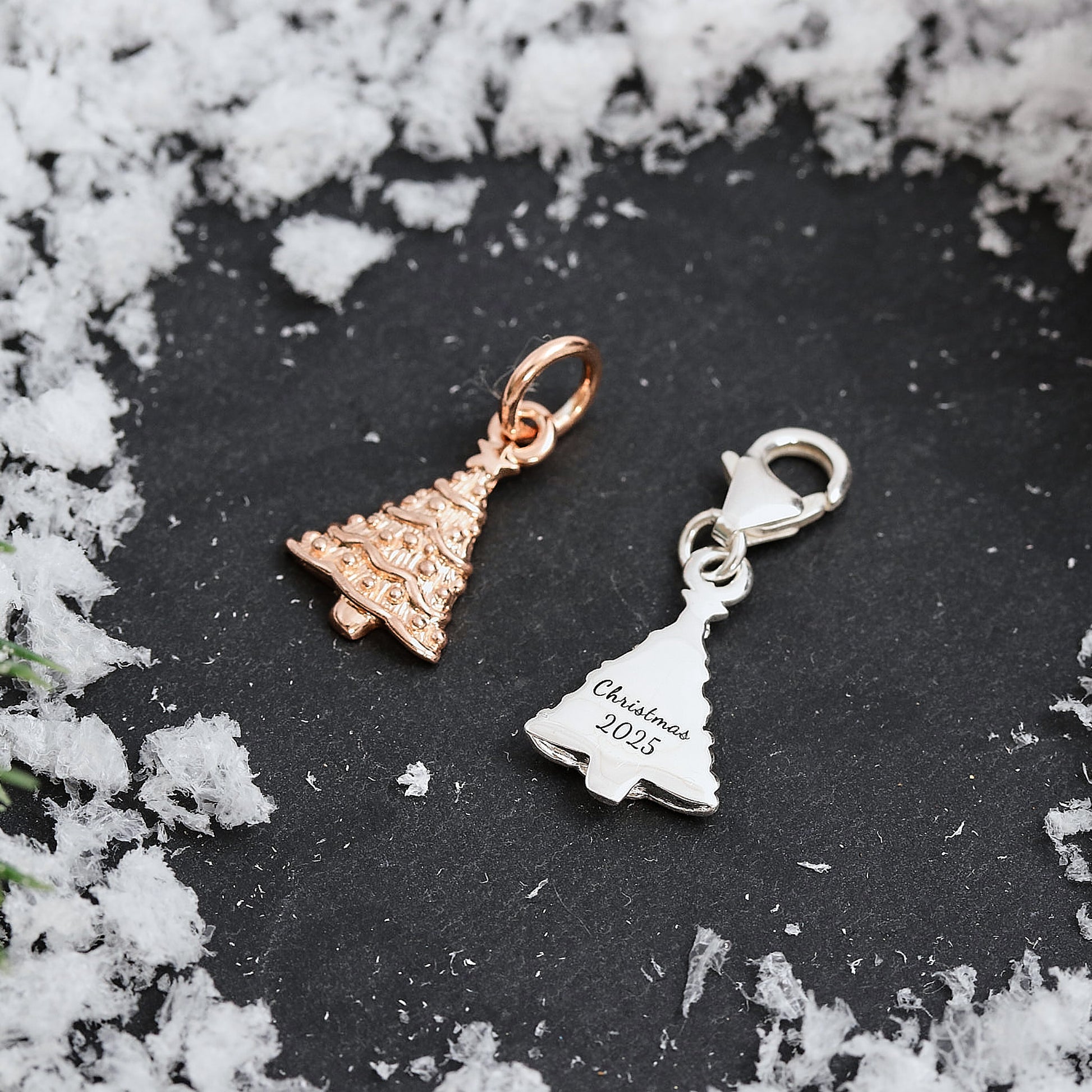 Two Christmas tree-shaped charms, one rose gold and one silver, on a dark surface with snow around.