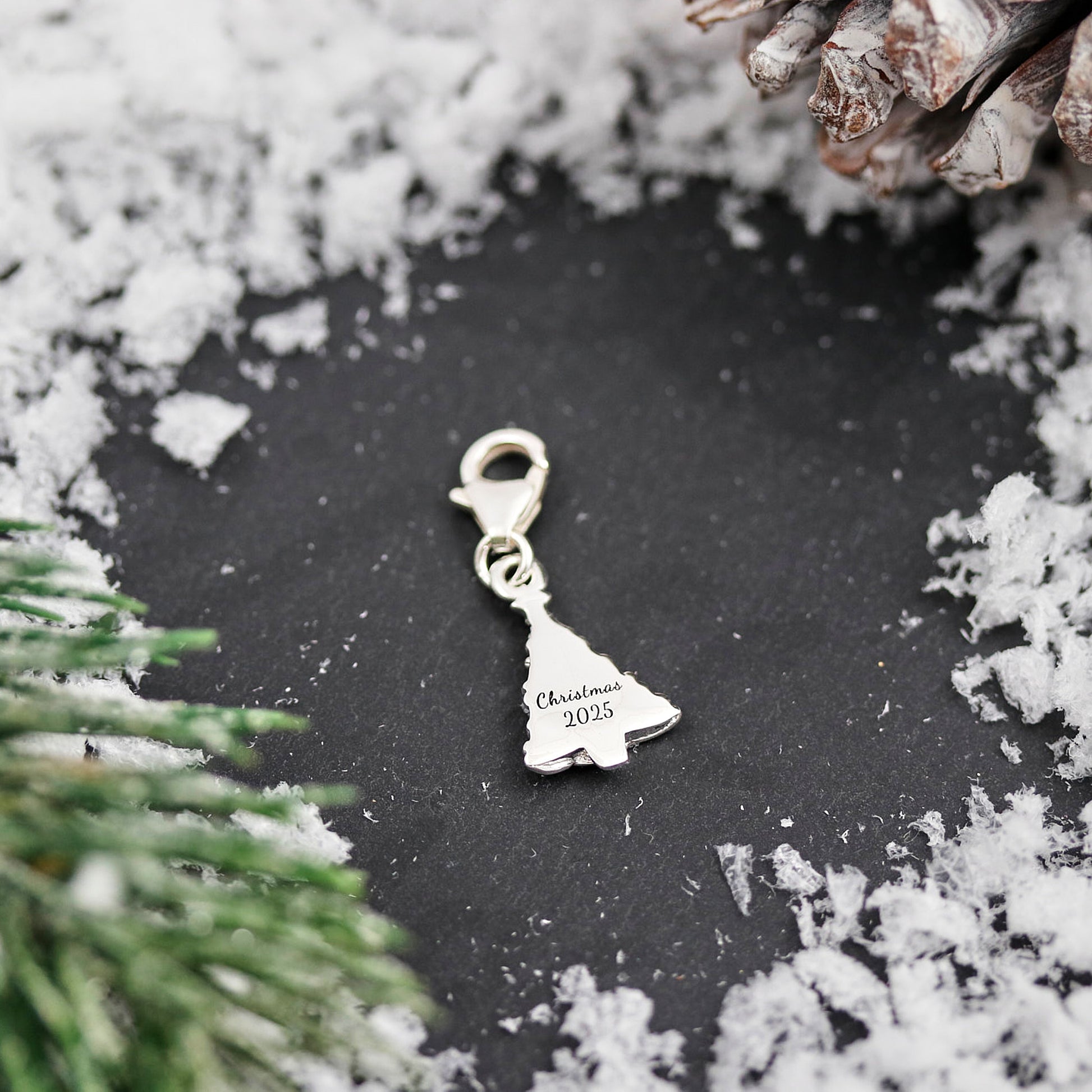 Silver Christmas tree charm with 'Christmas 2025' text on a black background with snow and pine branches.