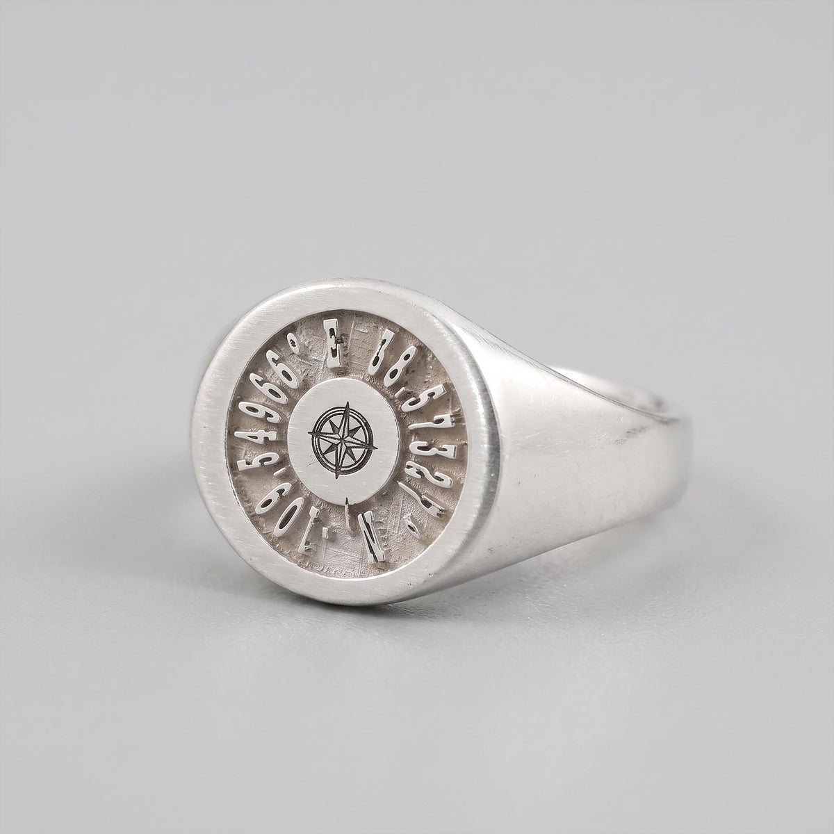 A silver signet ring  featuring coordinates and a compass rose, shown on a light gray background 