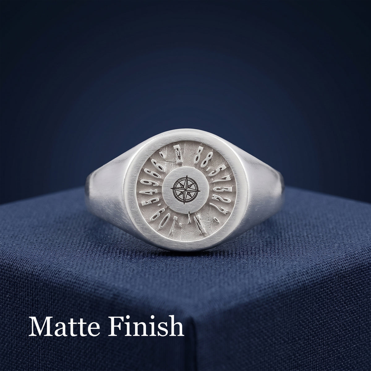 A silver signet ring with a matte finish, featuring a circular face engraved with coordinates and a compass rose in the center, resting on a textured navy blue surface with 'Matte Finish' text overlay.
