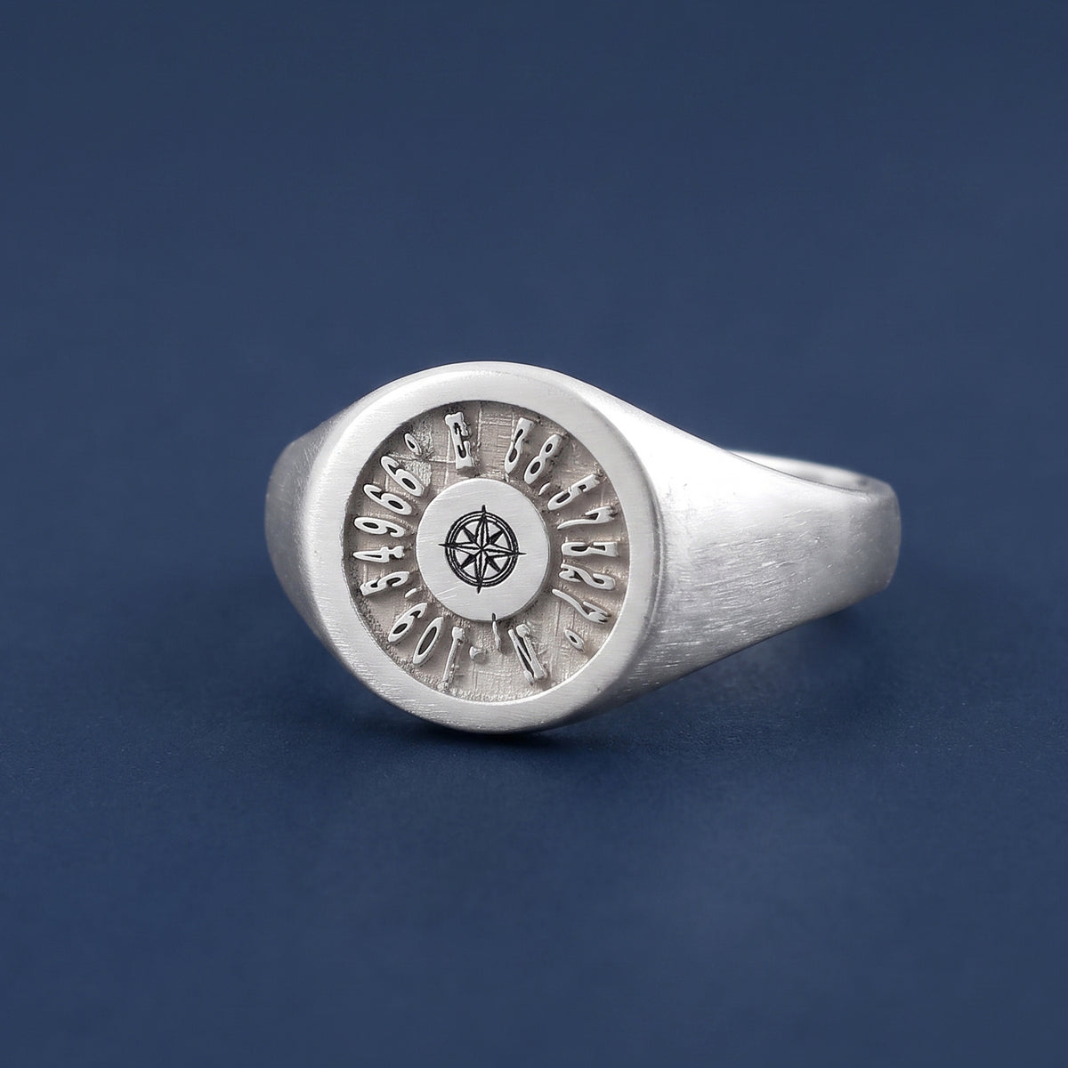 A silver signet ring featuring an engraved coordinate location or nautical chart design with a compass rose at the center, displayed on a dark blue background.