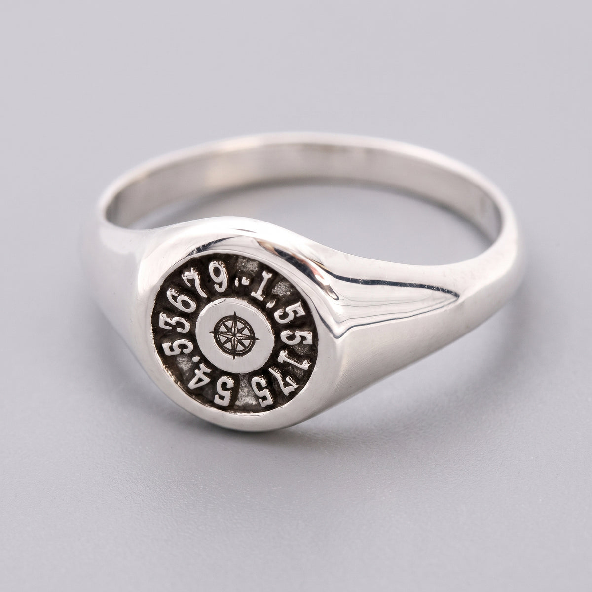 A sterling silver signet ring featuring a circular face engraved with a compass rose and latitude longitude coordinates, shown on a neutral gray background.