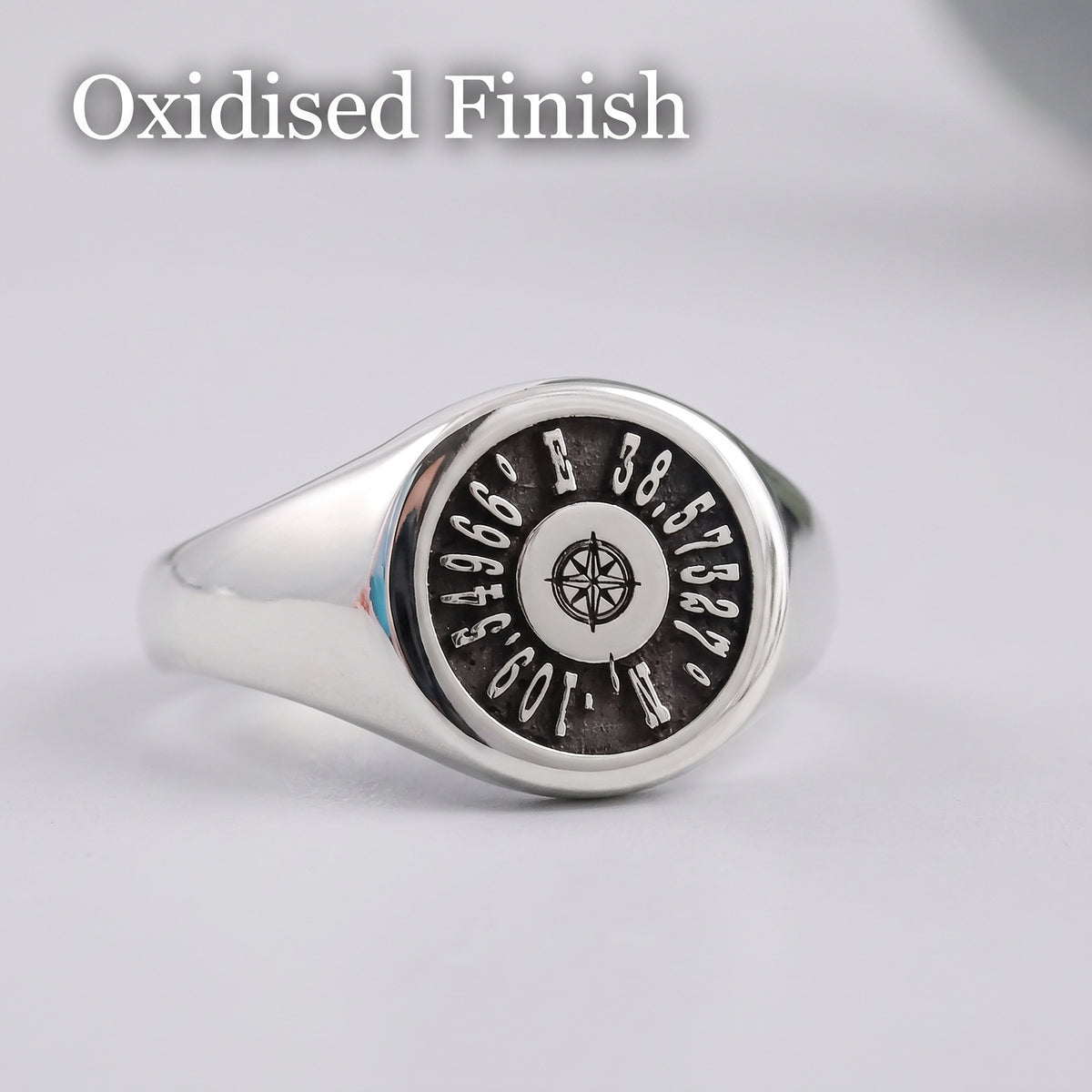 A silver signet ring with an oxidized black face featuring coordinates and a compass rose, shown on a light gray background with the text 'Oxidised Finish' at the top.