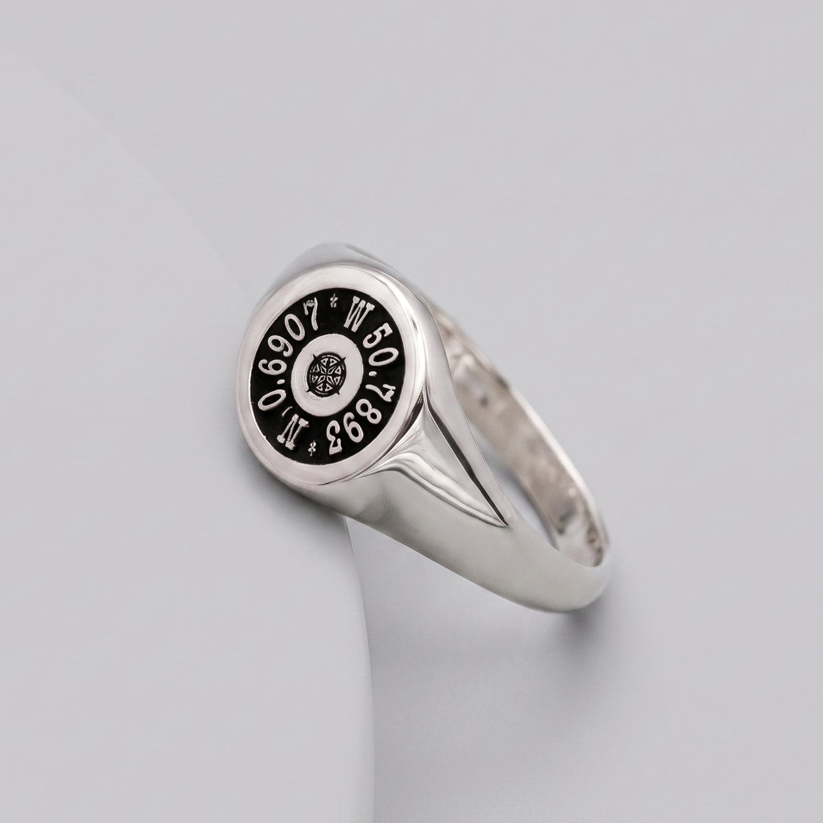 A silver signet ring with a black circular dial face featuring white nautical compass markings coordinates, shown on a light gray background.