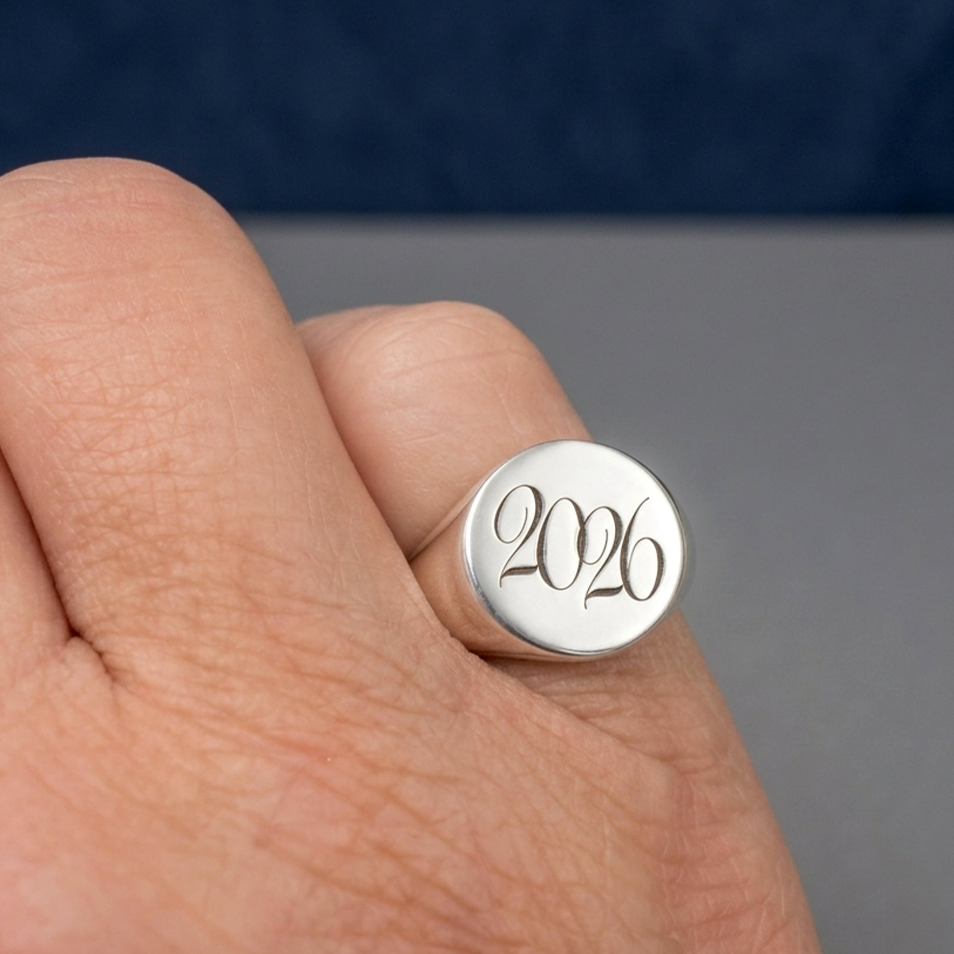 A close-up of a hand wearing a silver signet ring with the year '2026' engraved on the flat circular face.