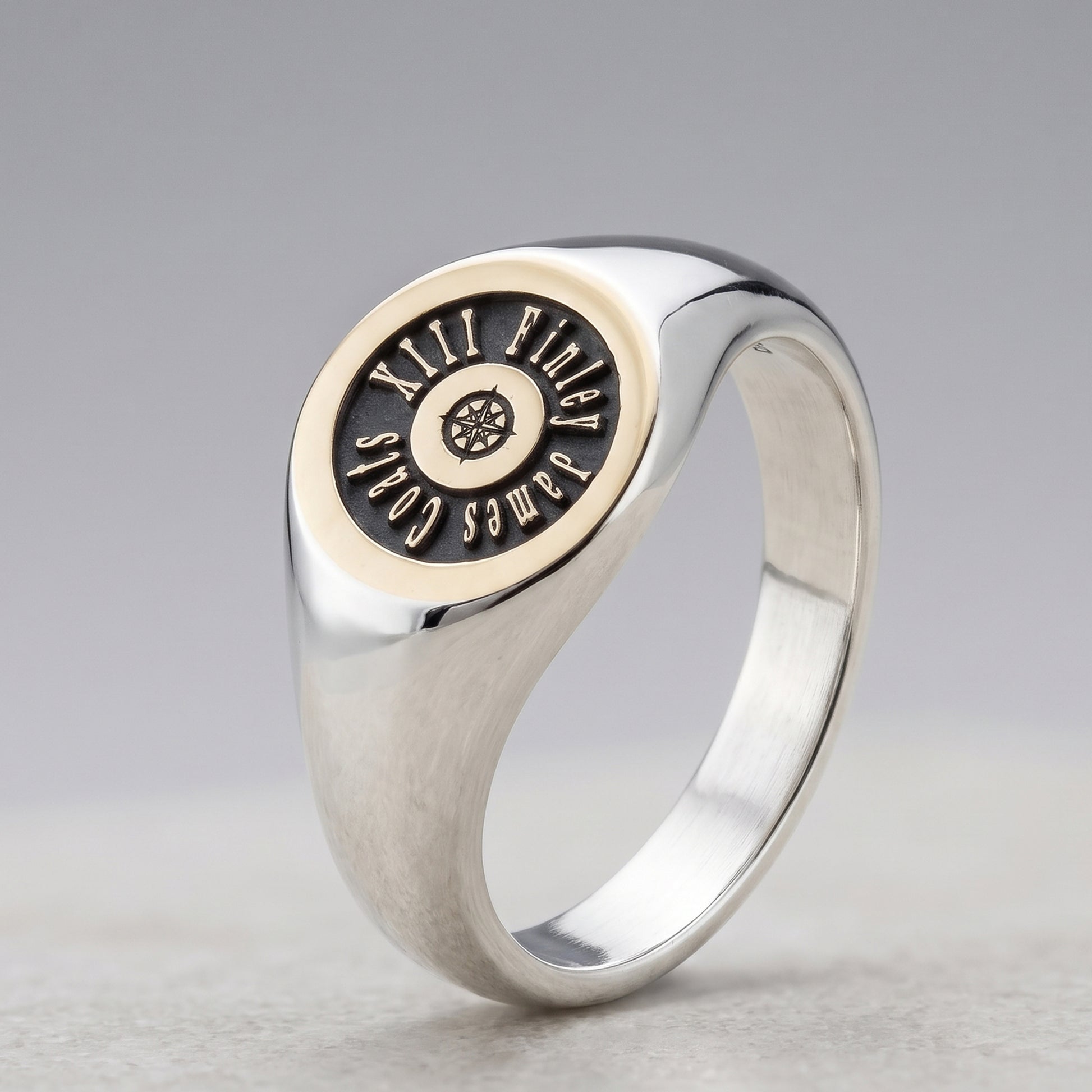 A silver signet ring with a circular face featuring a black inlay lettering that reads 'JAMES COLES'.