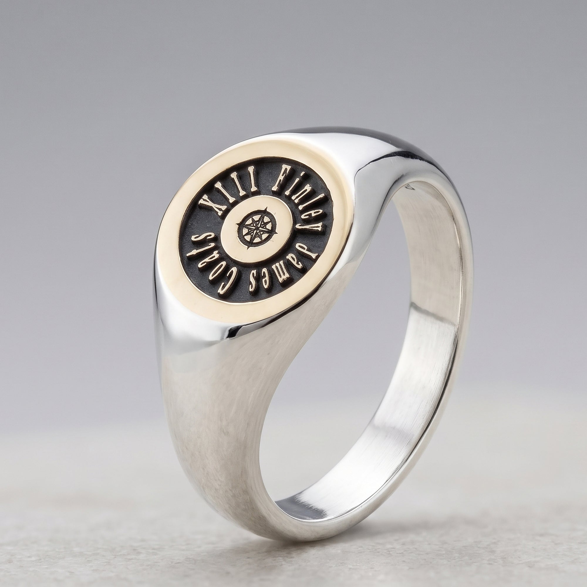 A silver signet ring with a circular face featuring a black inlay lettering that reads 'JAMES COLES'.