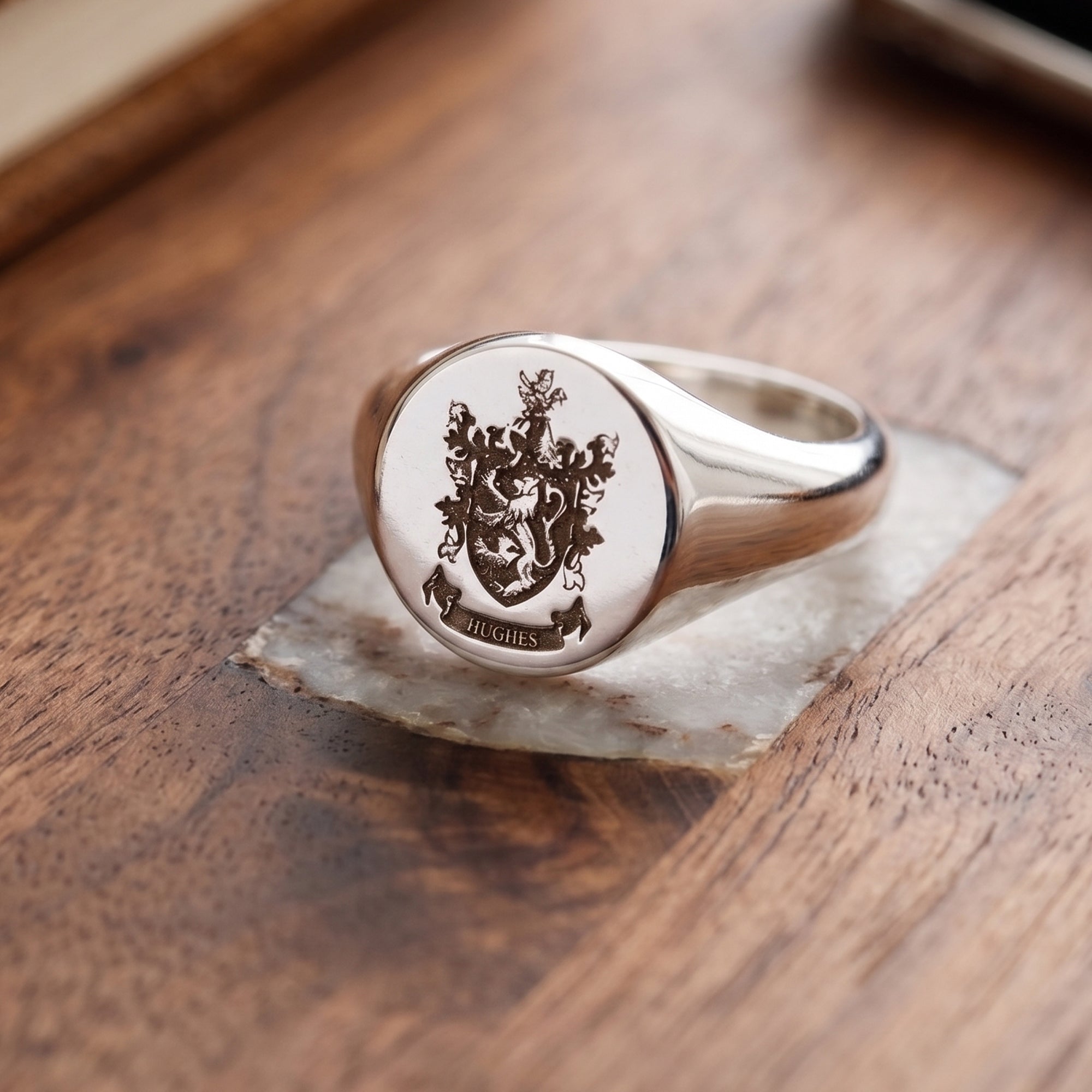 A silver signet ring with a detailed crest and the name 'HUGHES' engraved on the face, resting on a small piece of white marble over a wooden surface.