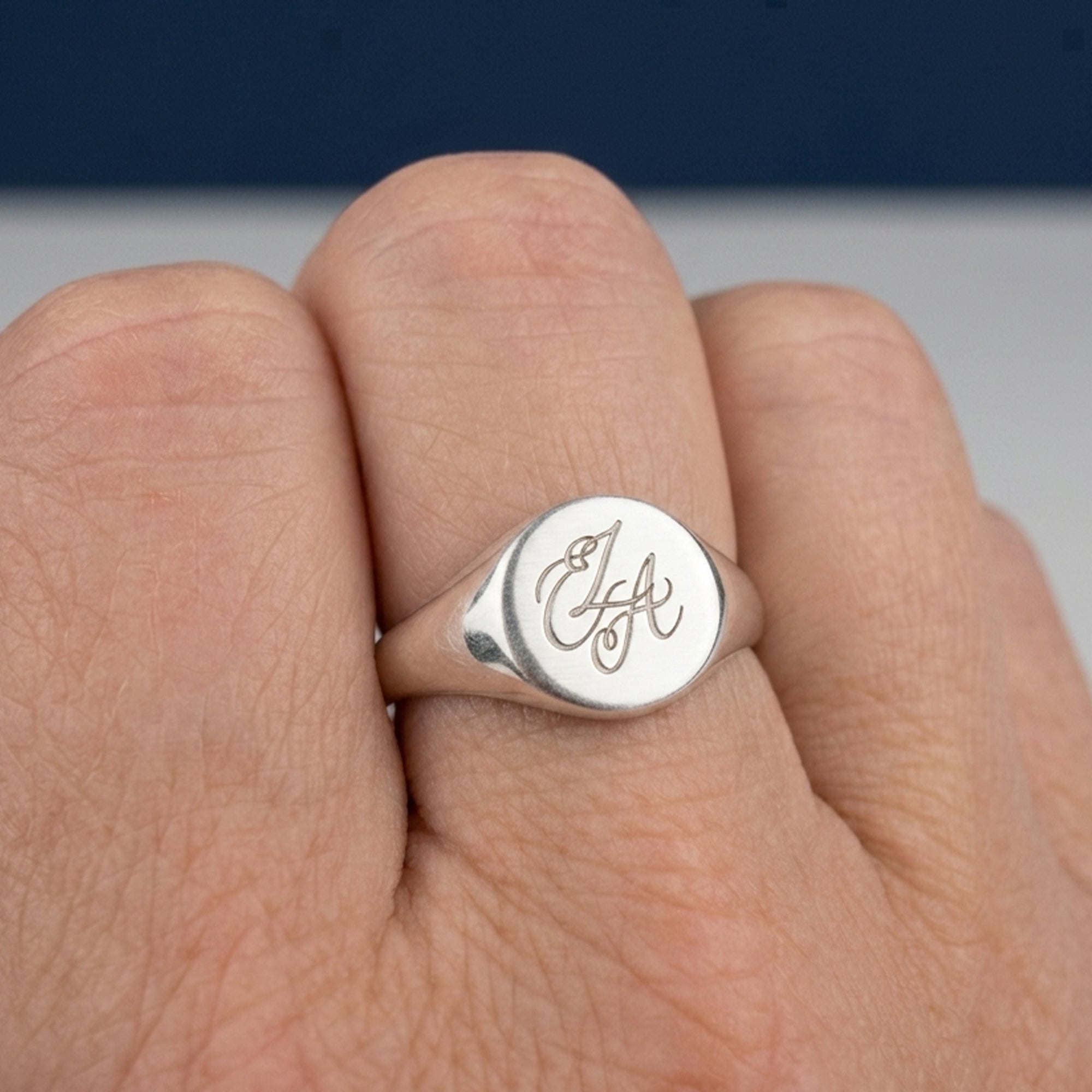 A close-up of a silver signet ring with an engraved cursive monogram on a finger, set against a blurred background.