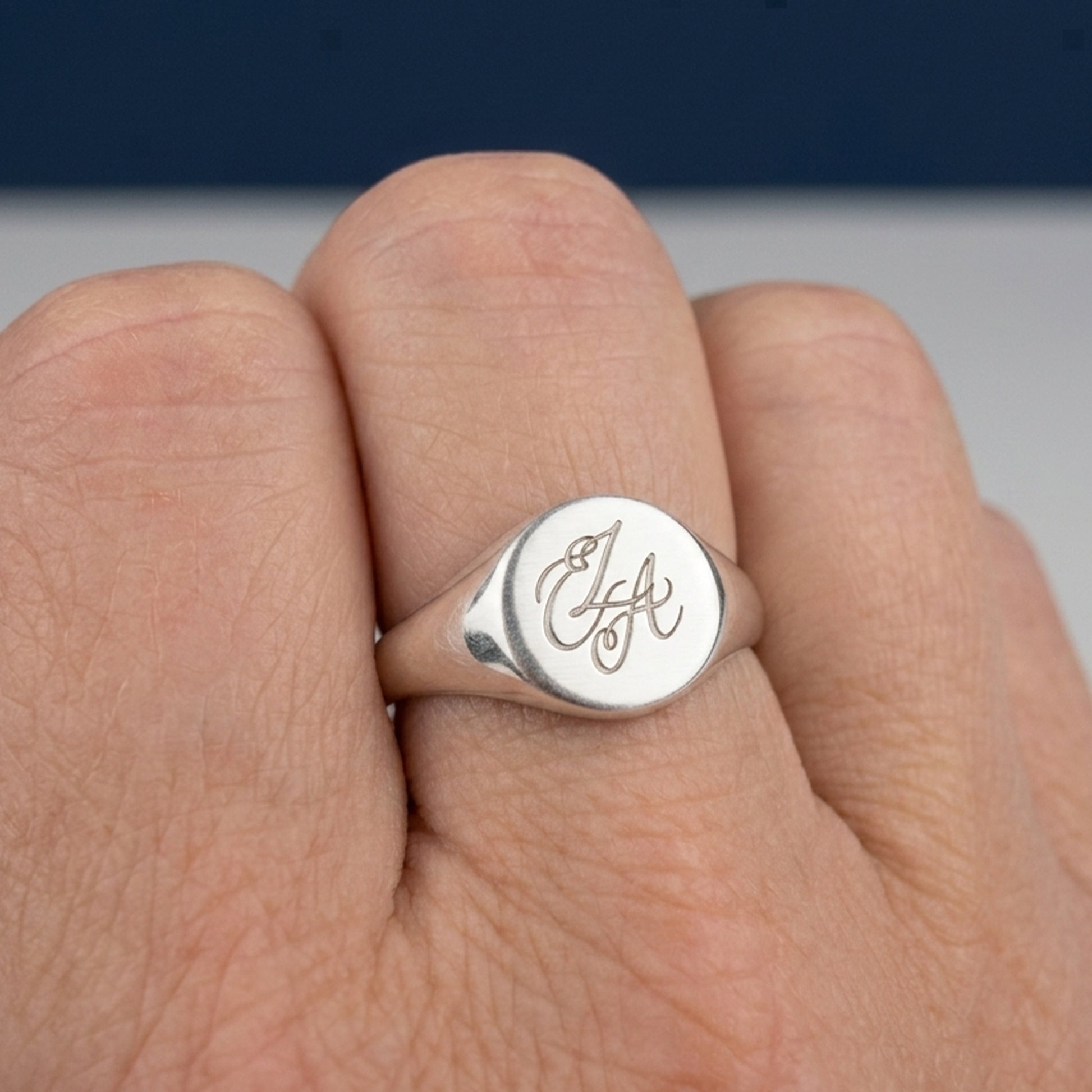 A close-up of a silver signet ring with an engraved cursive monogram on a finger, set against a blurred background.