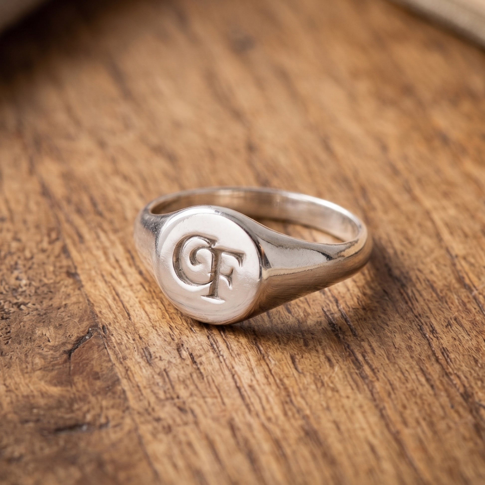 A polished silver signet ring with a circular face engraved with the initials 'CF', resting on a textured wooden surface.