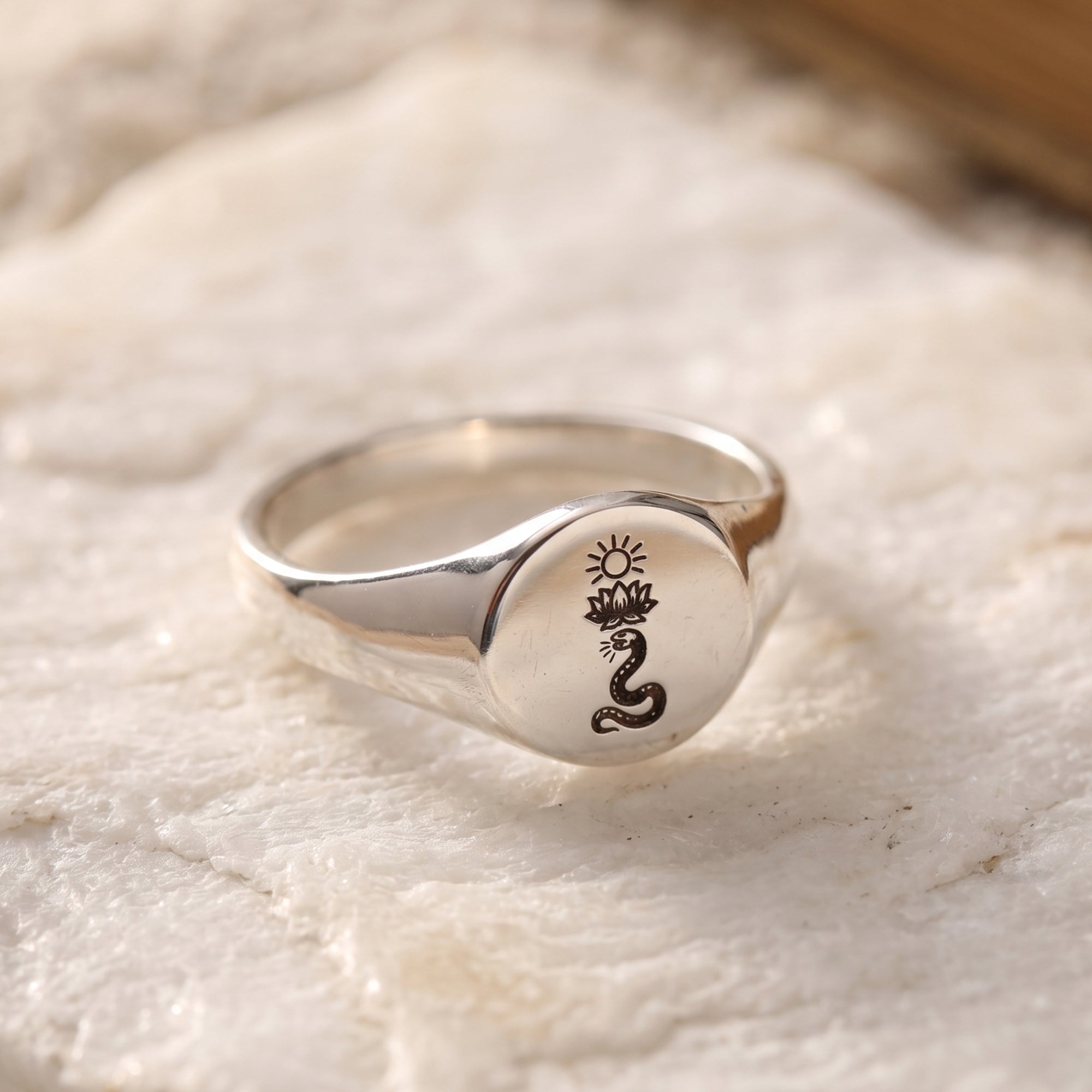 A polished silver signet ring with a black engraved lotus and sun motif, resting on a textured white stone surface.
