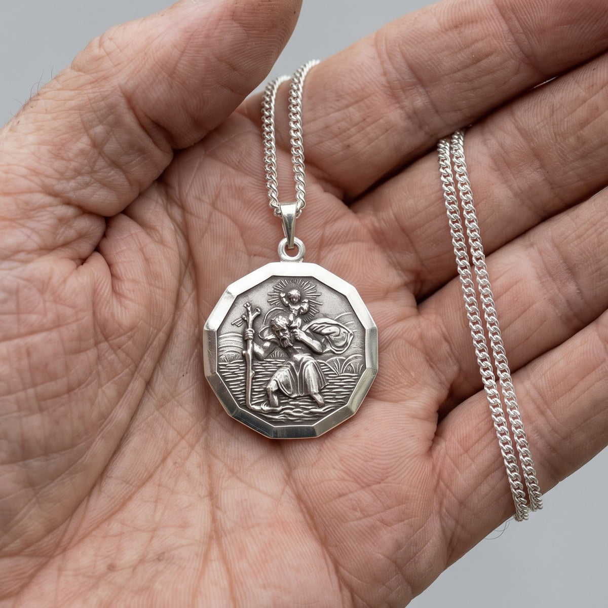 A silver-toned octagonal pendant necklace featuring a detailed relief carving of Saint Patrick, displayed resting on an open hand. The pendant is suspended from a silver curb chain, with the chain draped over the fingers of the hand against a neutral gray background.