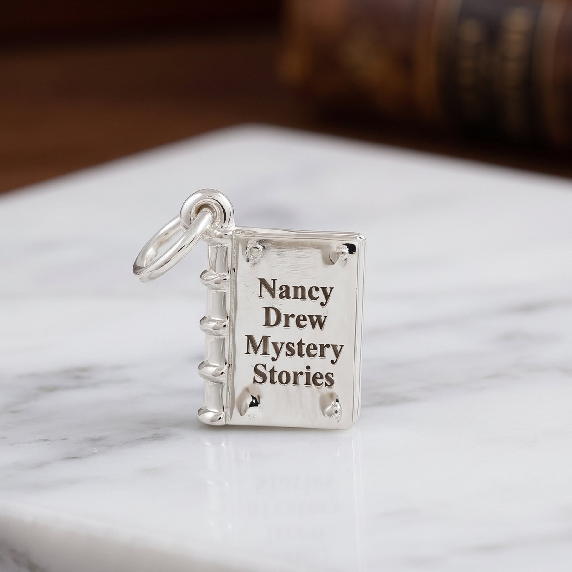 A silver book-shaped pendant charm engraved with the text 'Nancy Drew Mystery Stories' and attached to a silver ring, resting on a white marble surface.