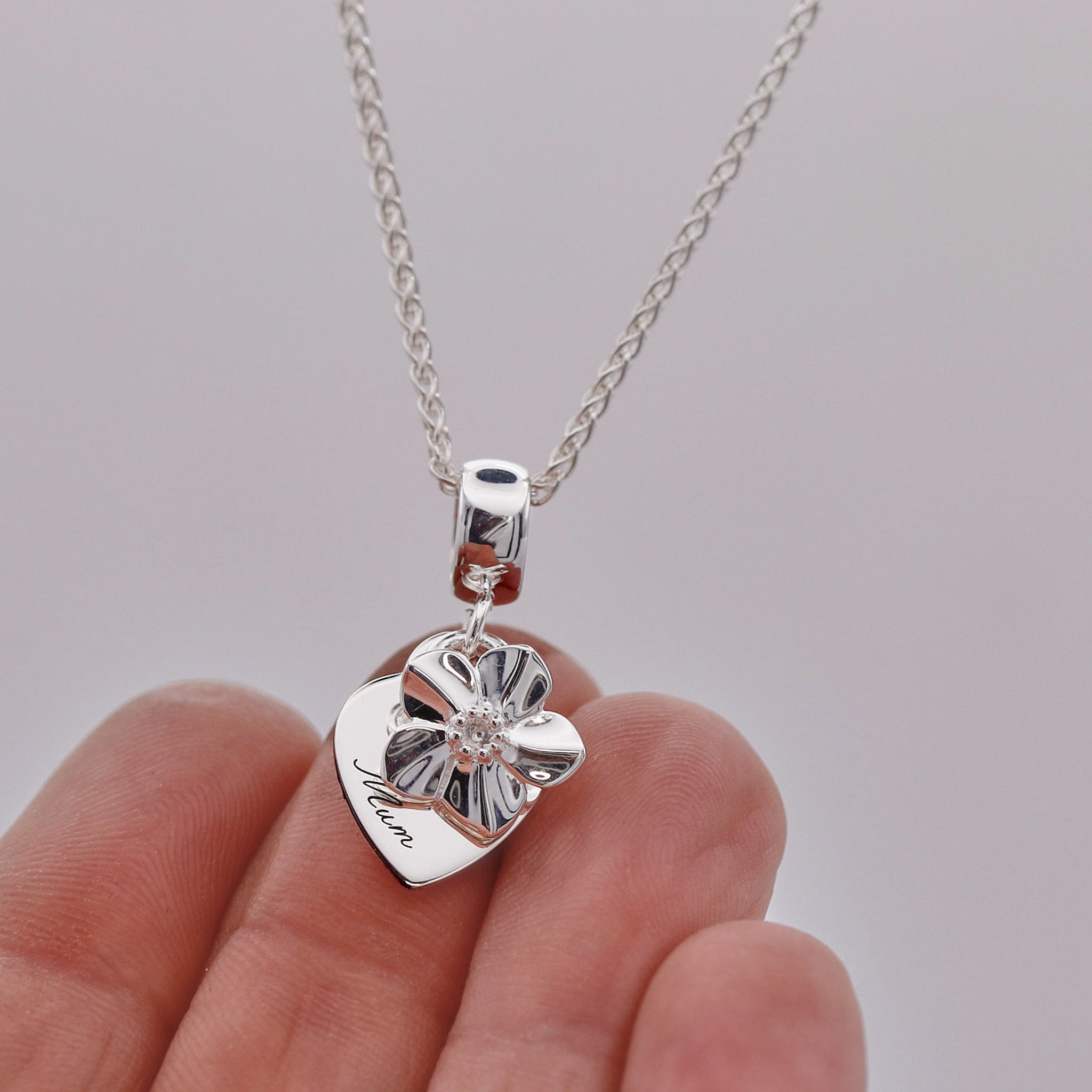 Silver heart-shaped pendant with 'Mum' engraved on a chain held by a hand against a gray background