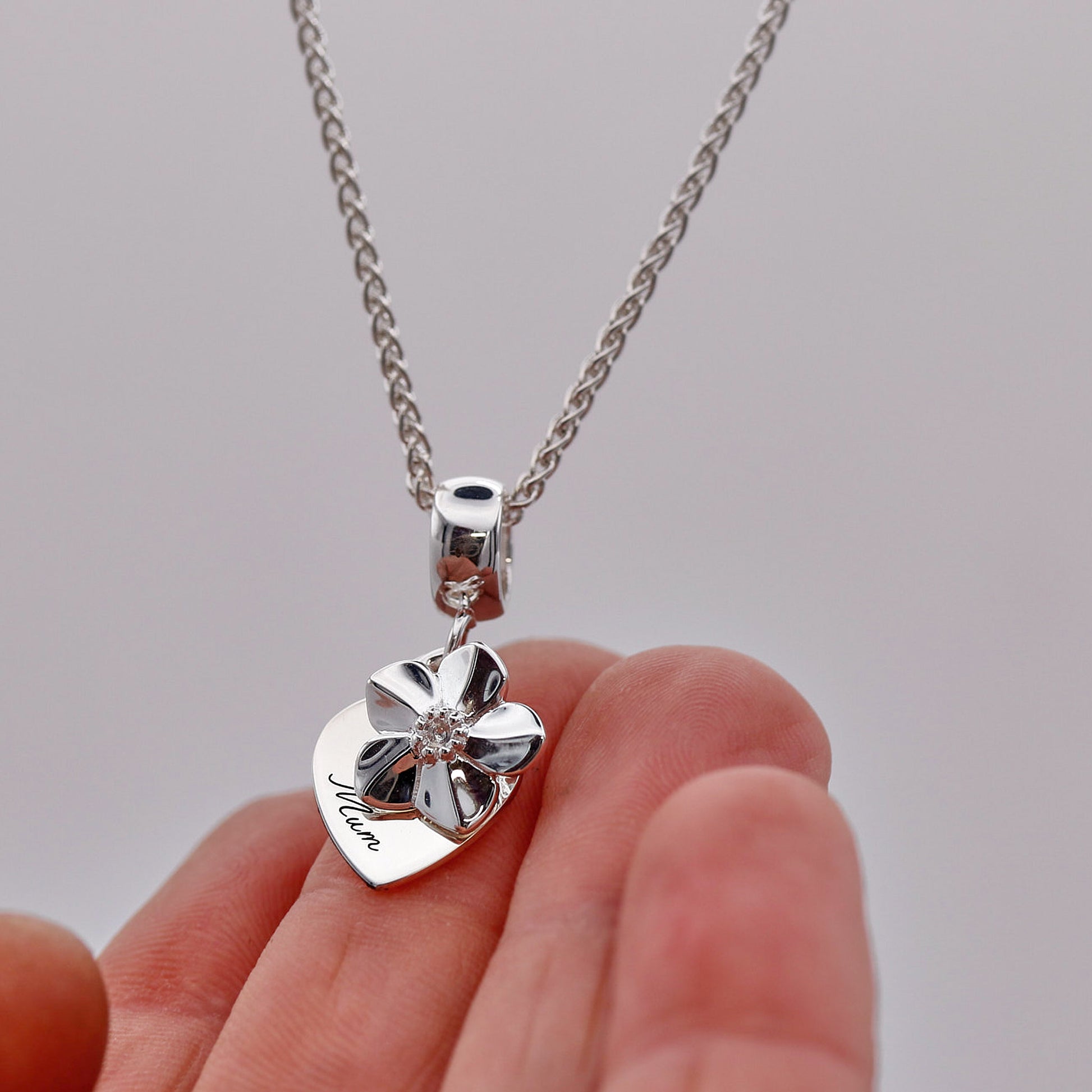 Silver heart-shaped pendant with 'Mum' engraving on a chain held by a hand against a gray background
