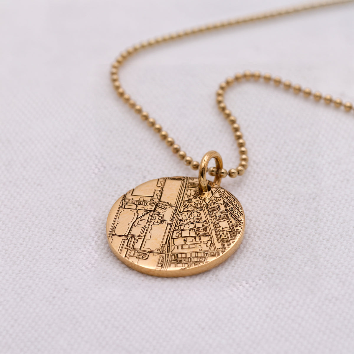 A gold necklace with a round, engraved map pendant on a matching ball chain, displayed on a textured white fabric background.