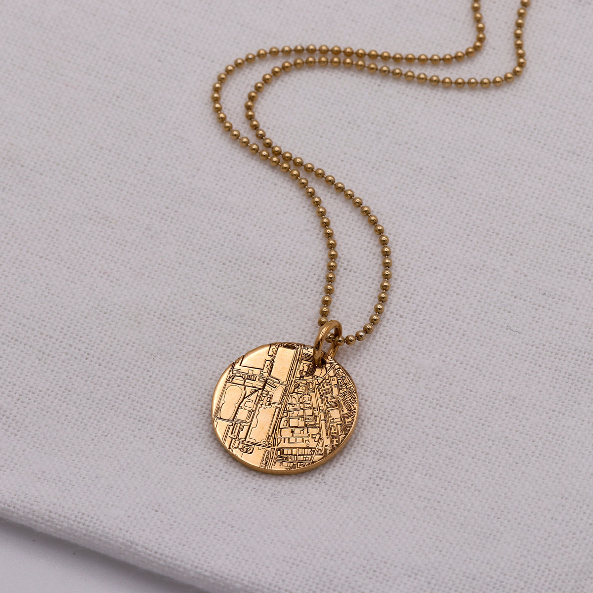 A gold-tone necklace with a round map-engraved pendant on a gold ball chain, displayed on a textured white fabric background.
