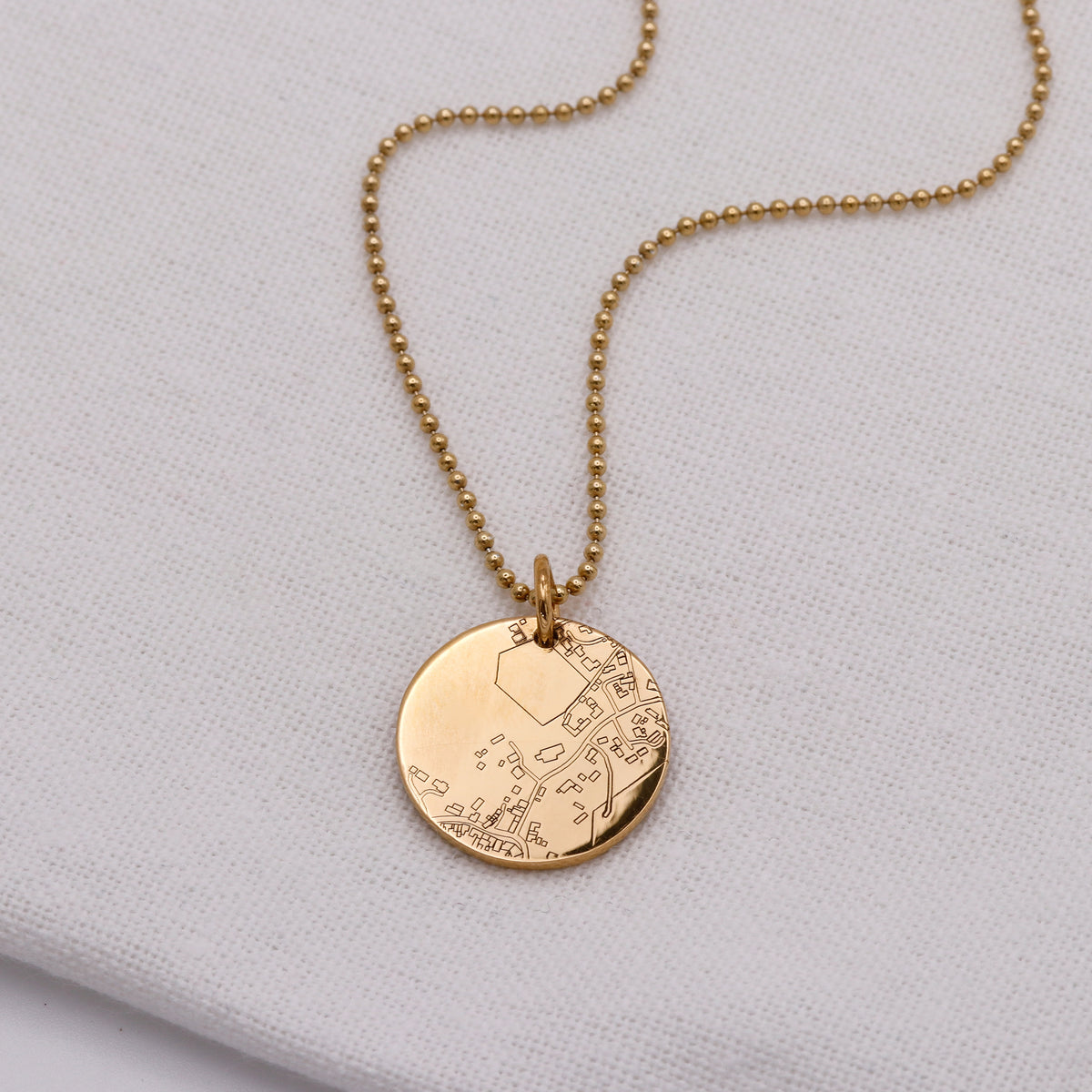 A gold-tone ball chain necklace with a round engraved pendant, displayed on a textured white fabric background.